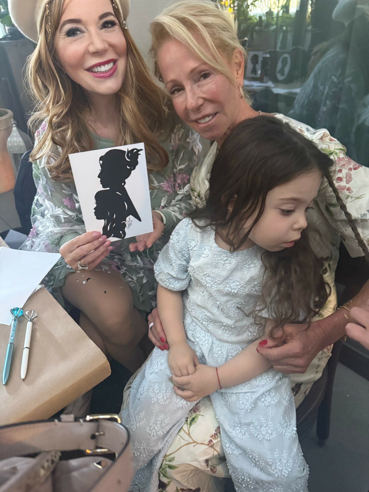 Three women, one holding a silhouette card, sitting together outdoors, smiling and engaging with a young girl in a white dress.