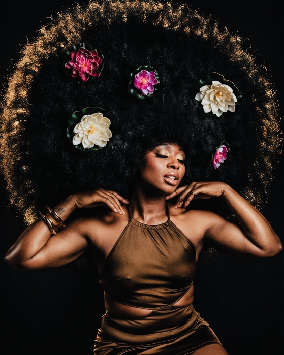 Woman with large afro hairstyle decorated with colorful flowers, wearing a brown sleeveless top, posing against black background.