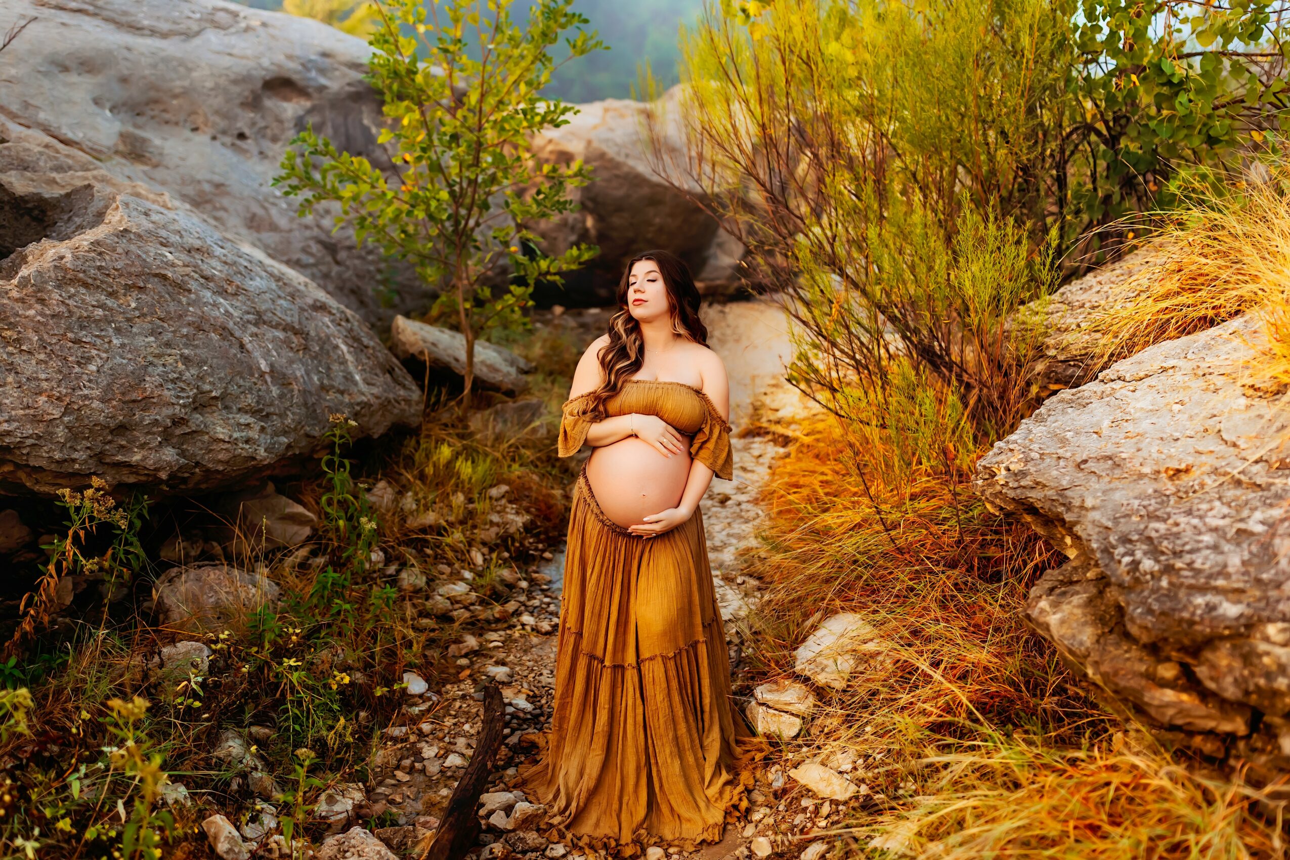 Pregnant woman in a long, flowing dress stands outdoors among rocks and bushes, holding her belly with both hands.