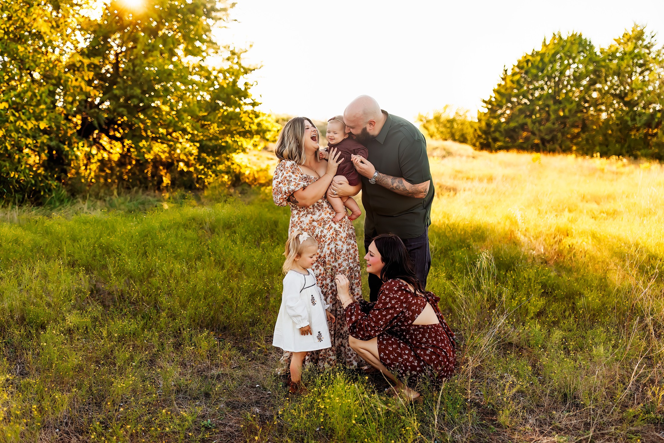 Family of four outdoors in a grassy field with trees, enjoying a sunny day, smiling and hugging each other.