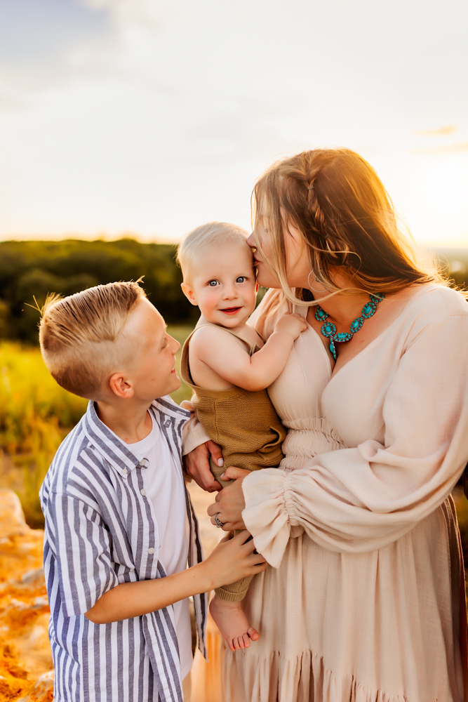 A woman holding a baby and a young boy outdoors during sunset, with a landscape background.