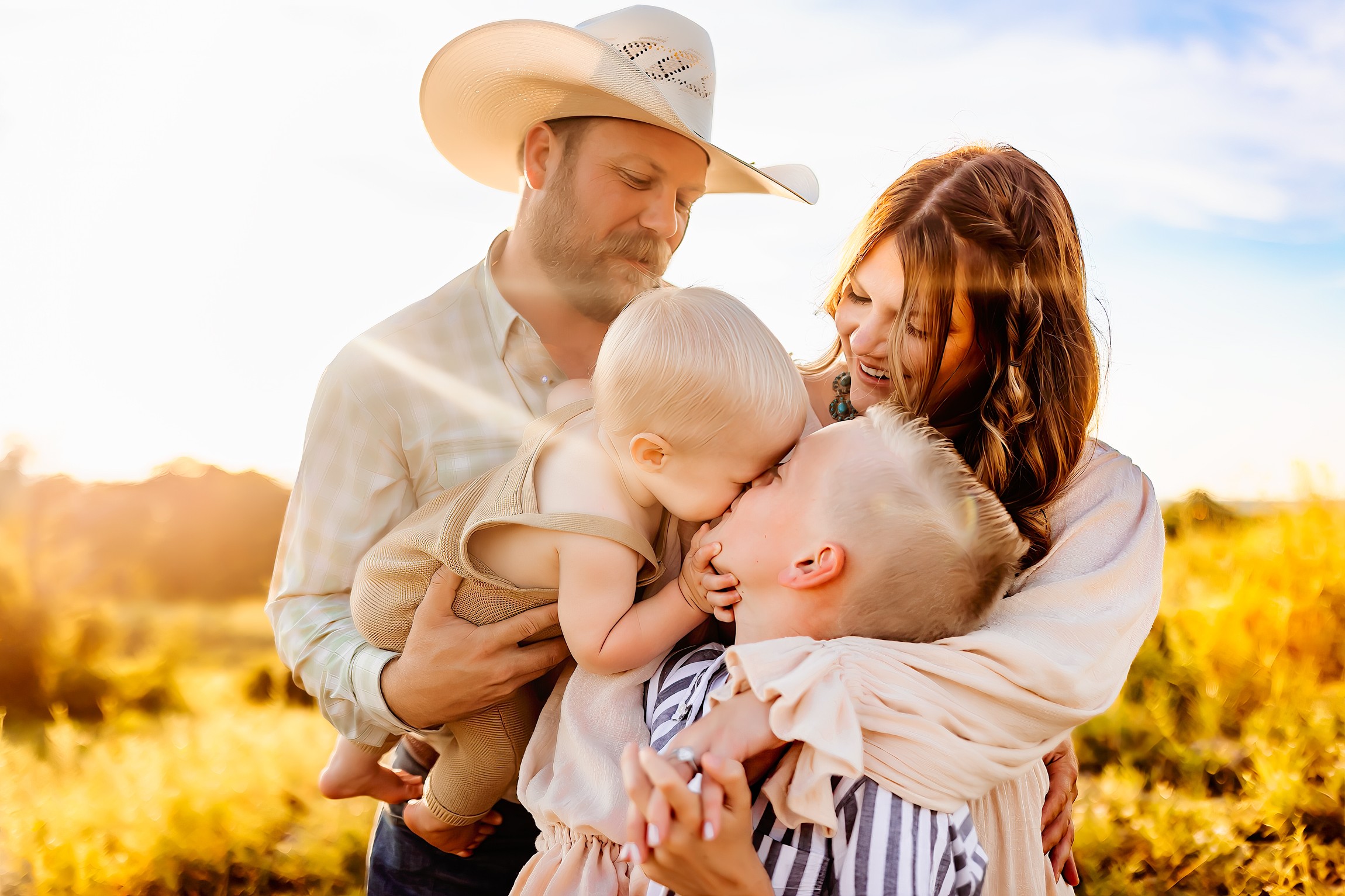 Family of four outdoors, parents holding two children, sunny sky, yellow field background