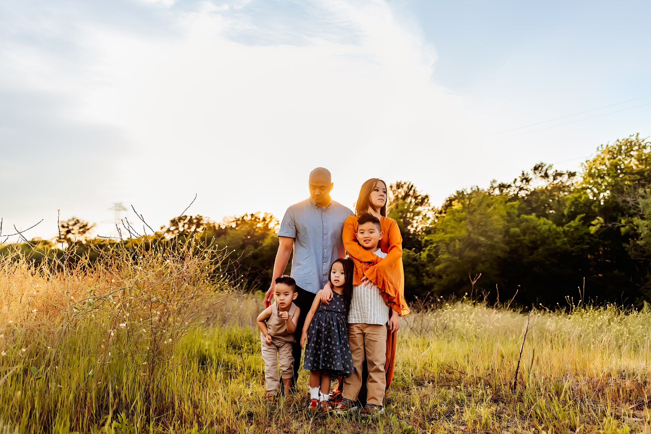 Family of five standing in a grassy field with trees and sky in the background, smiling and posing for a photo.