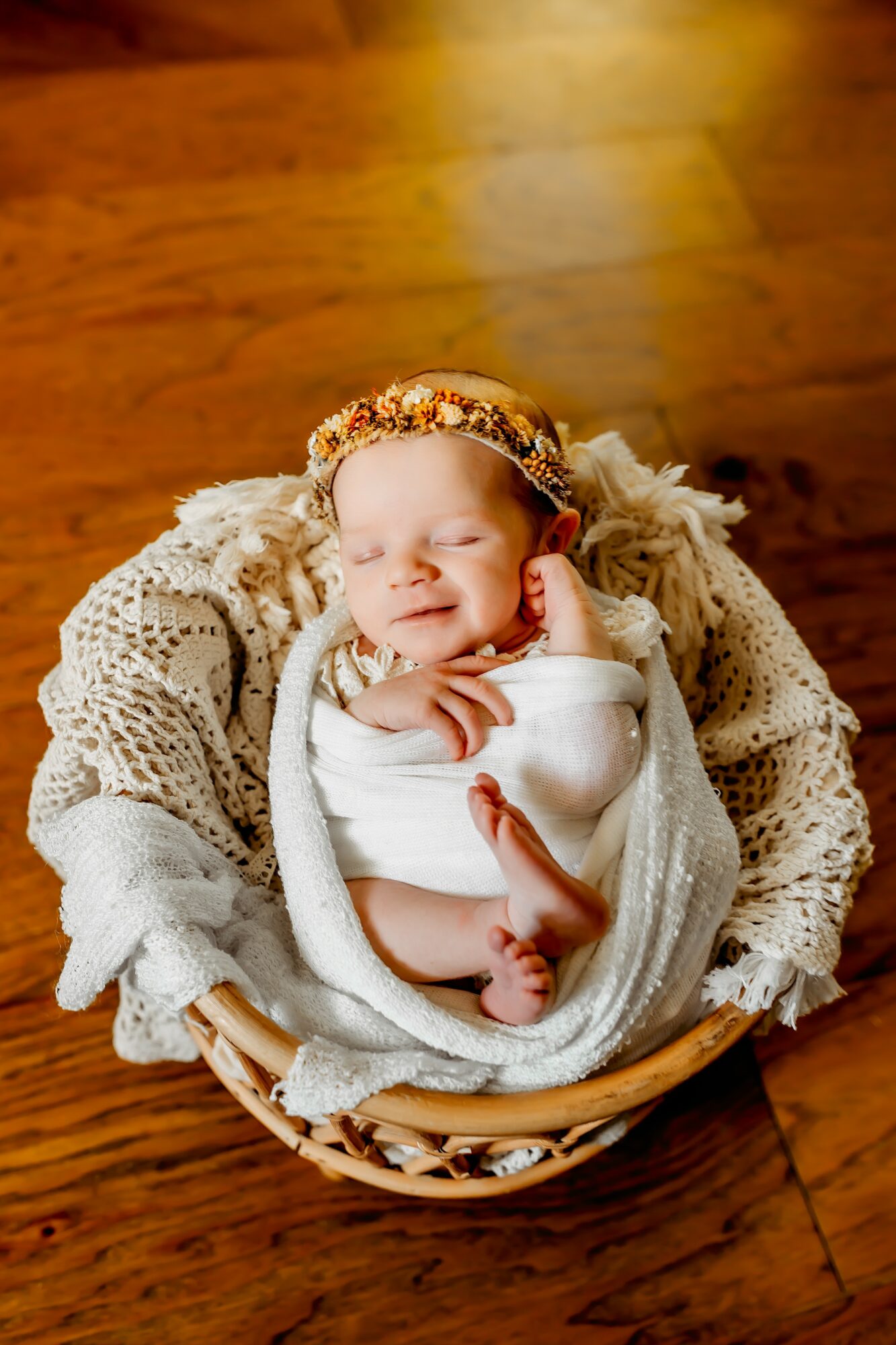 Baby with a floral headband, wrapped in a blanket, lying in a basket on a wooden floor.