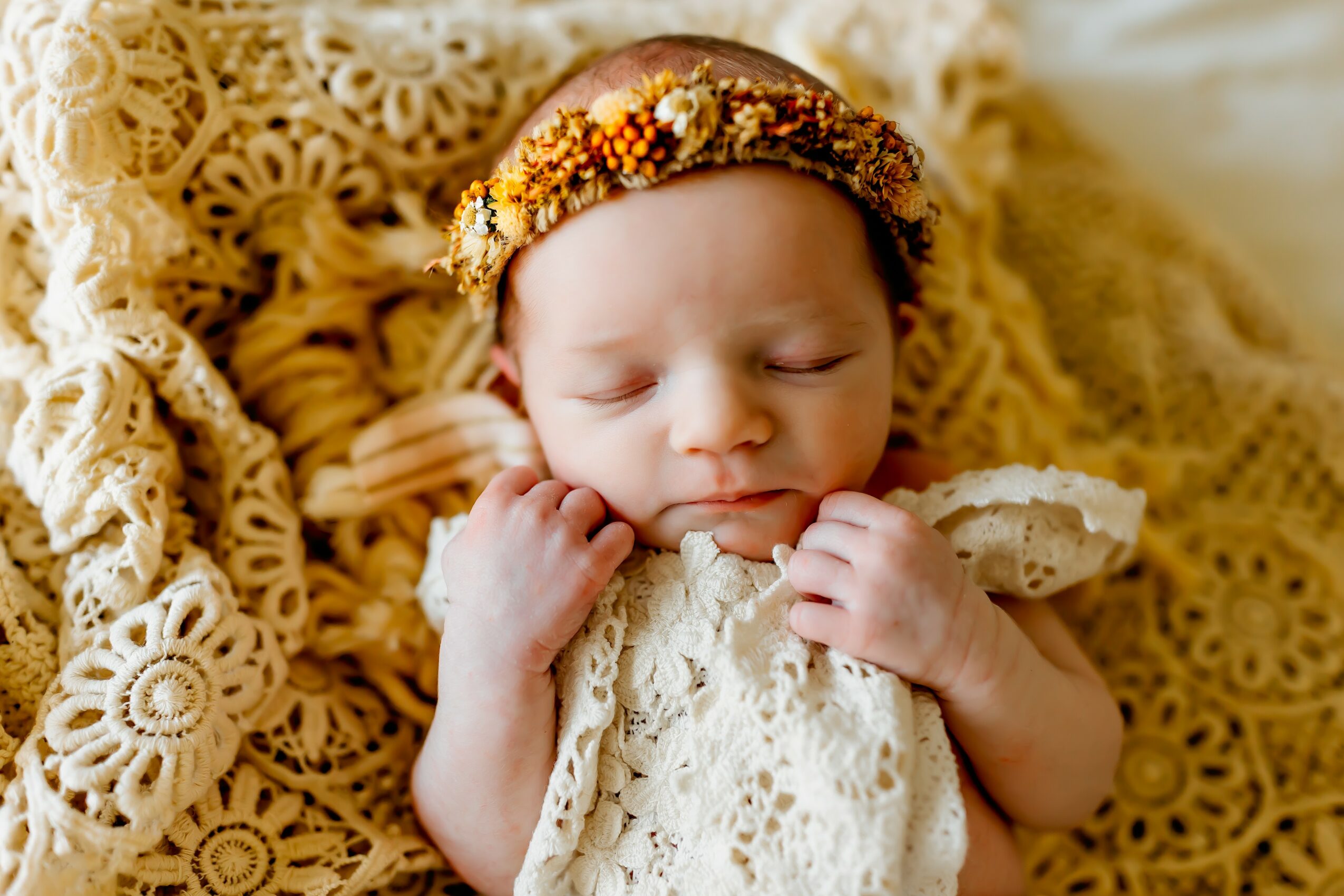 Baby with a floral headband, wrapped in lace, lying on a crocheted blanket, eyes closed, peaceful expression.
