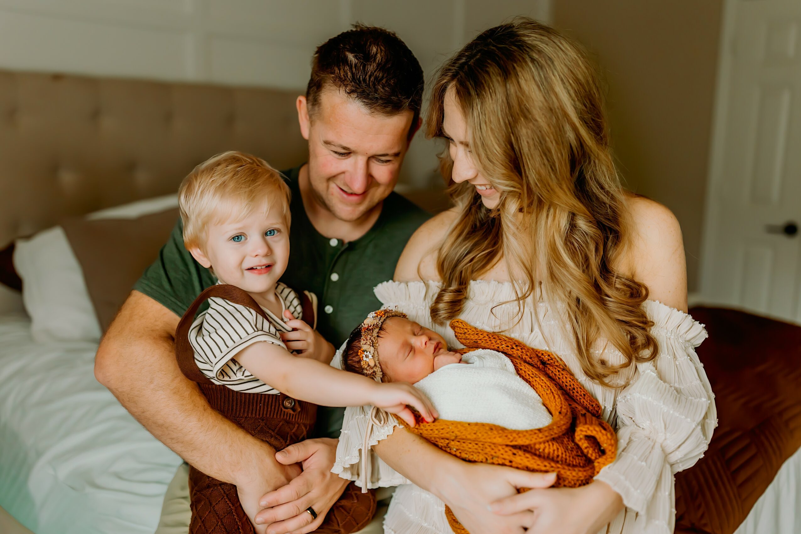 Family of four with a man, woman, young girl, and baby in a bedroom, smiling and interacting lovingly.