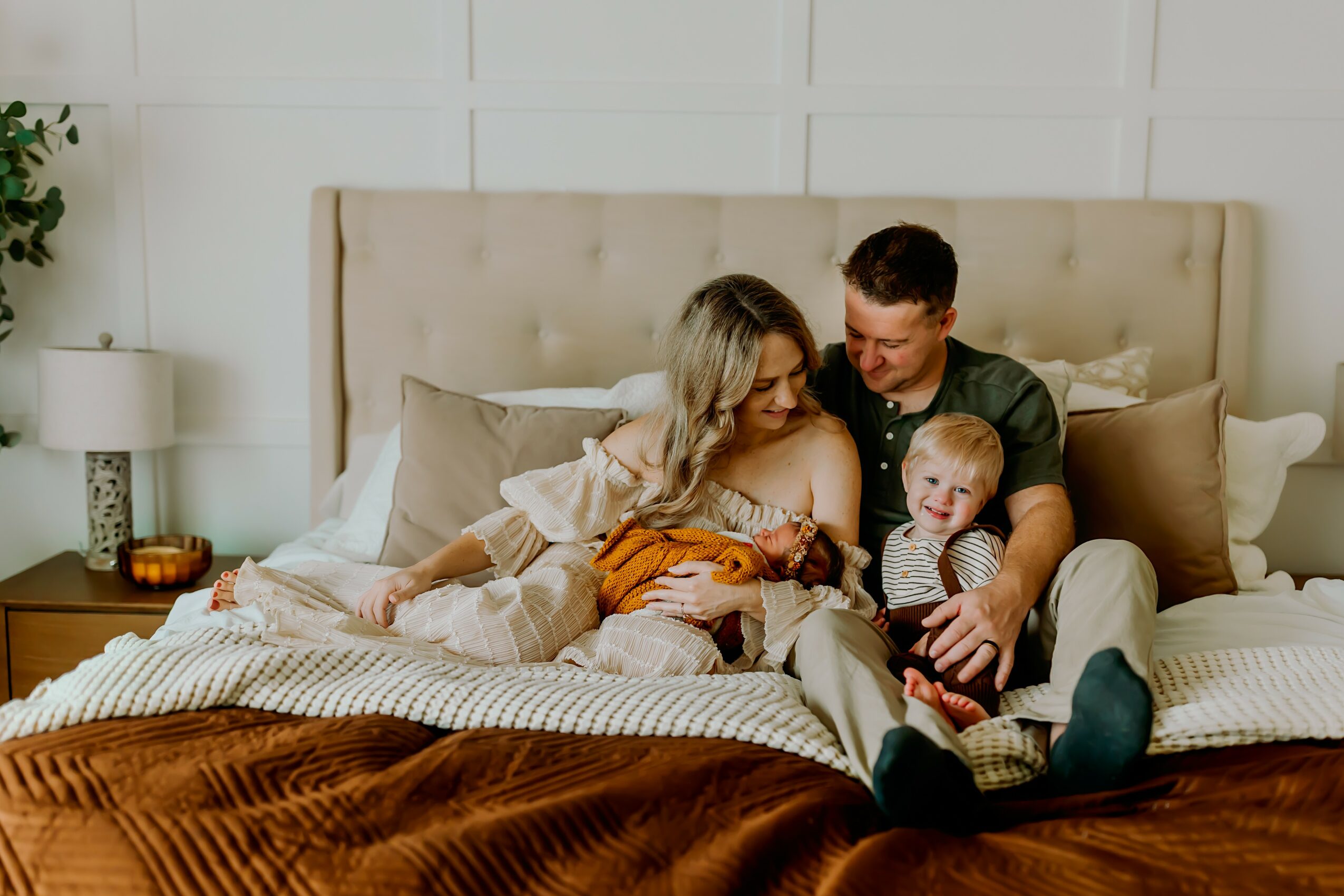 Family sitting on bed, smiling and looking at a tablet, in a bedroom with beige headboard and pillows.