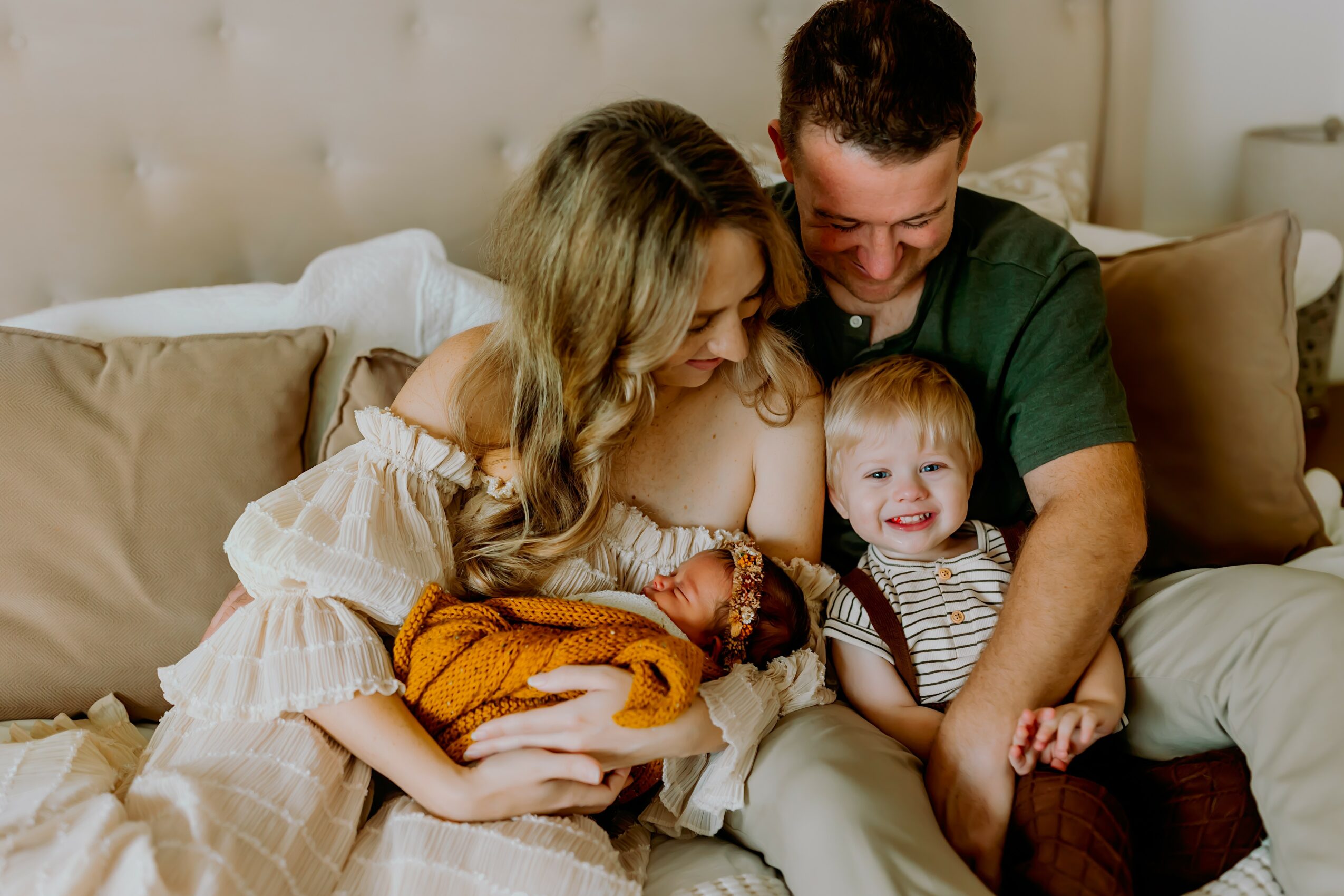 Family of four sitting on a bed, mother holding a newborn, father and young child smiling, in a cozy bedroom.