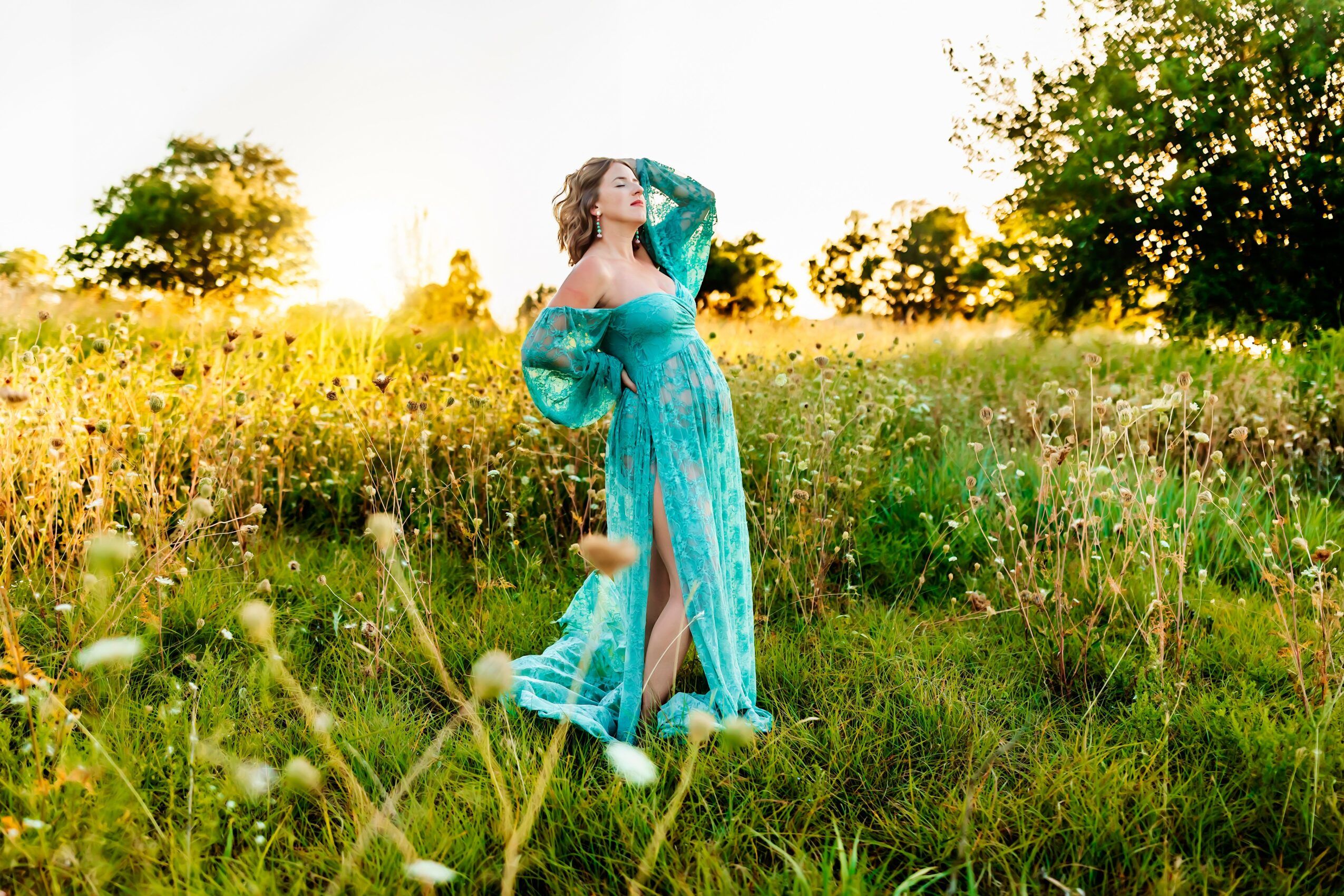 Woman in a flowing blue dress standing in a grassy field with trees and sunlight in the background.