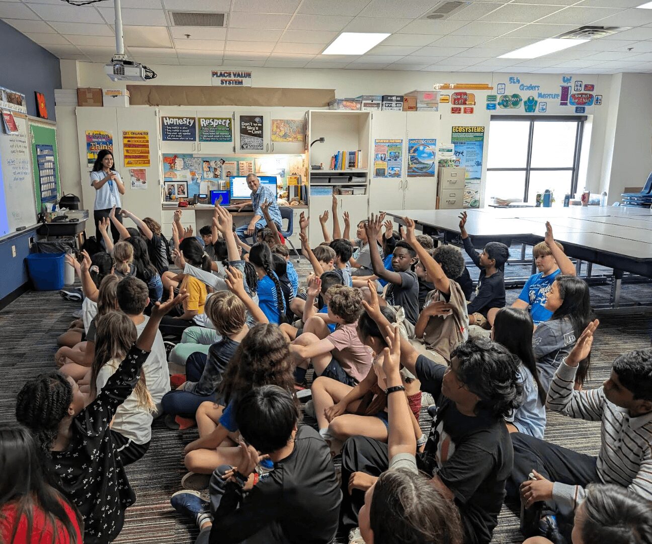 Classroom filled with children sitting on the floor, raising hands, with a teacher at the front near a whiteboard.