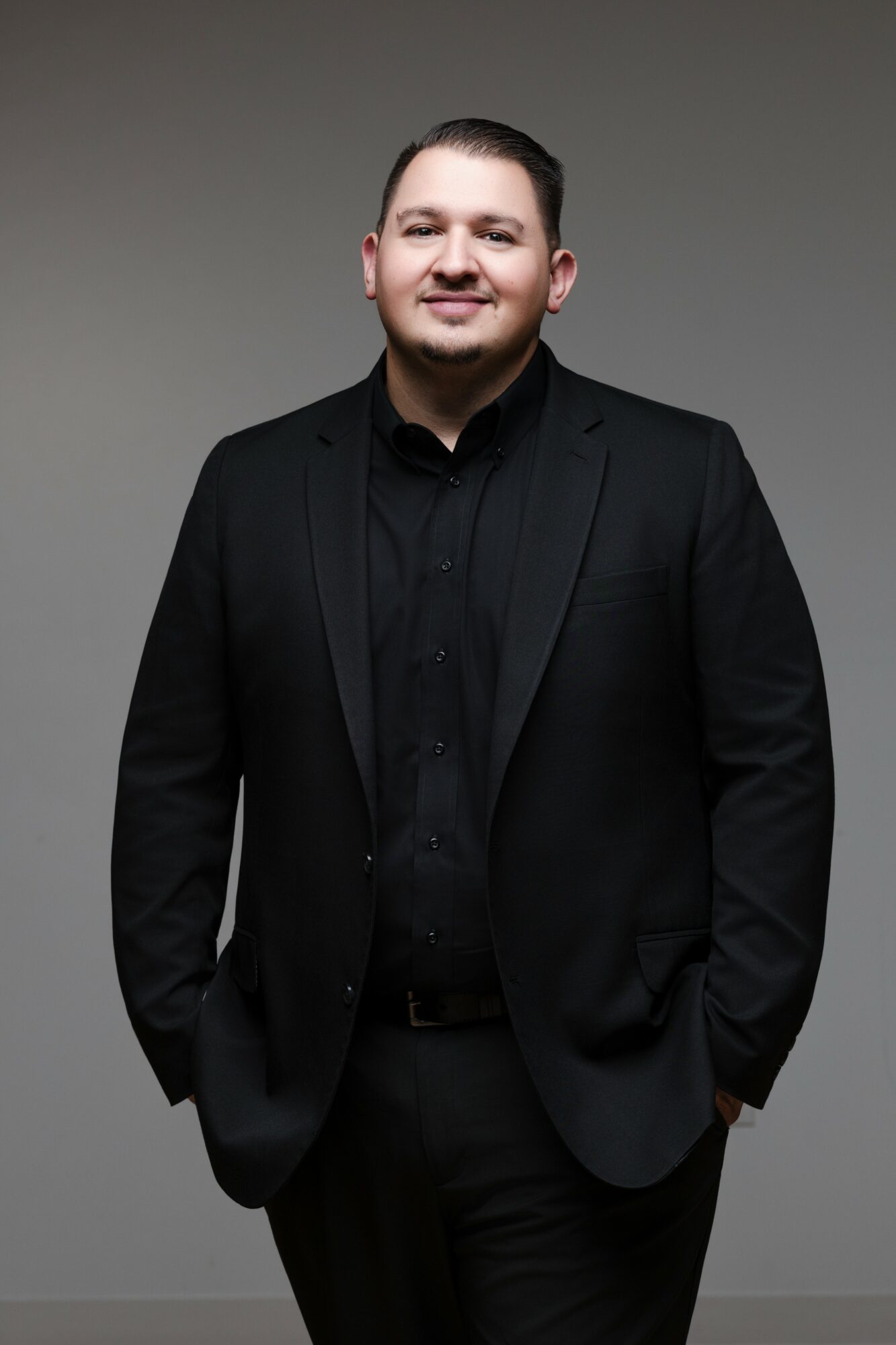 Man in black suit and shirt standing against a plain background, smiling, with hands in pockets.