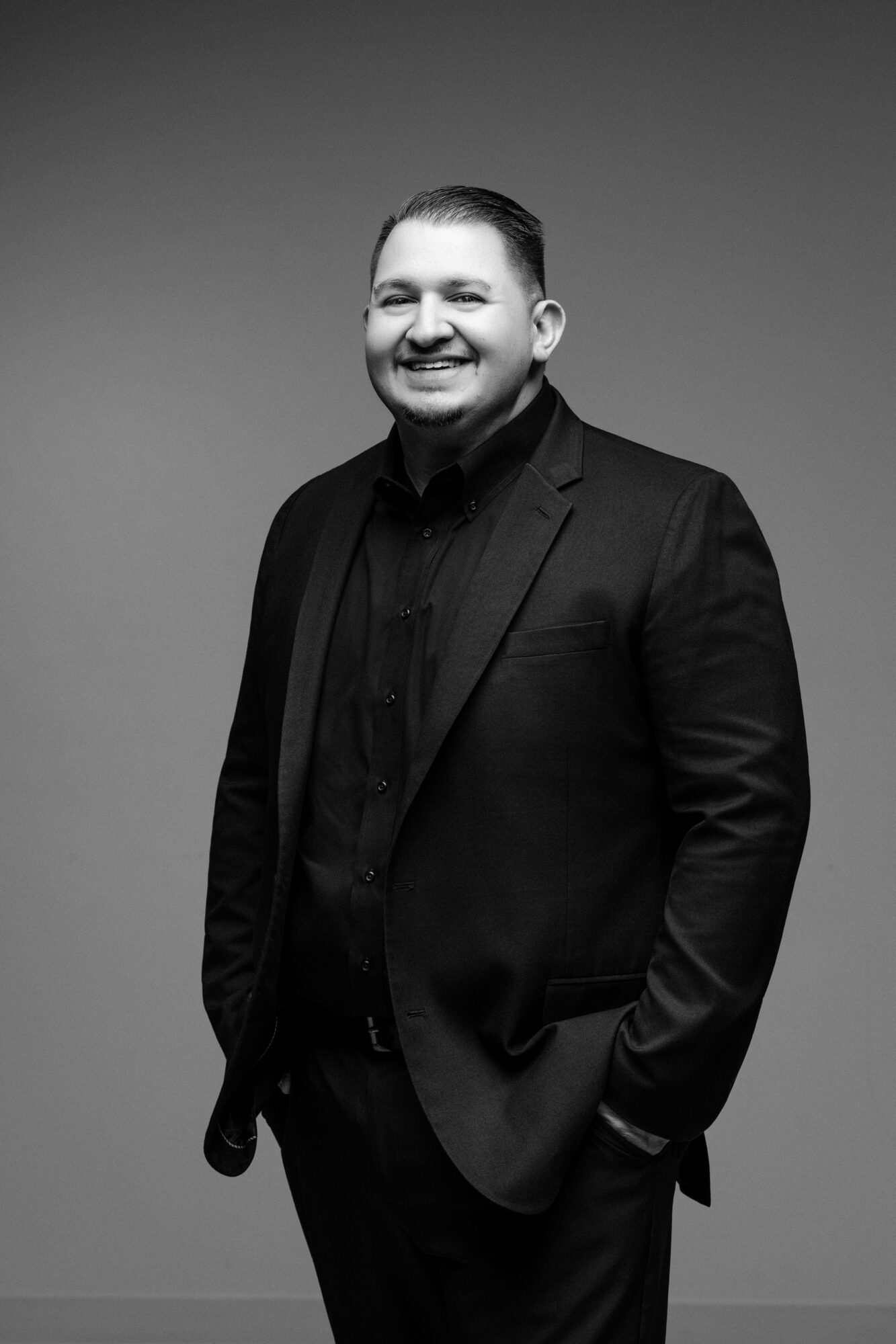 Man in suit smiling, standing with hands in pockets, against plain background.