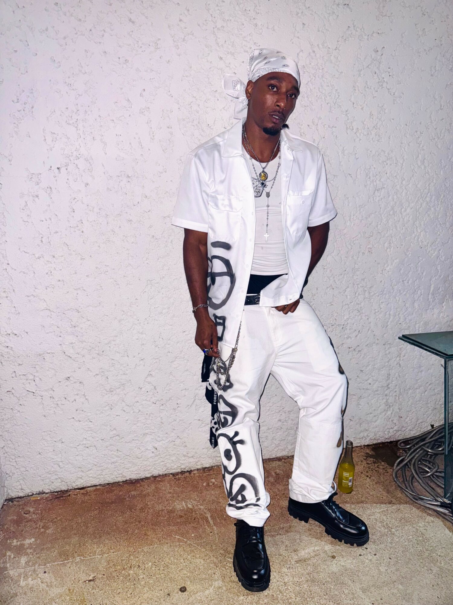 Young man in white outfit with black shoes, standing against a plain wall, with a small table nearby.