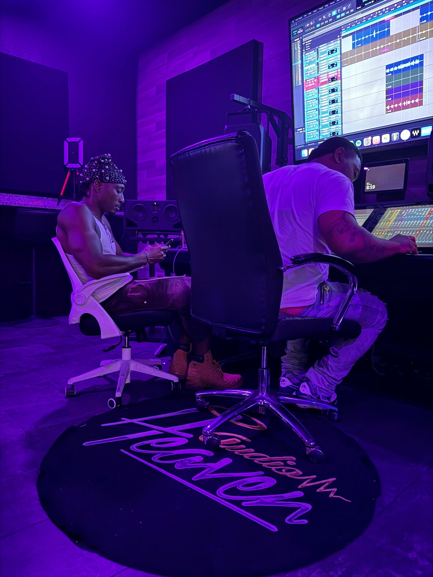 Two people in a music studio with a mixing console, purple lighting, and a round mat with text, one sitting and one standing.
