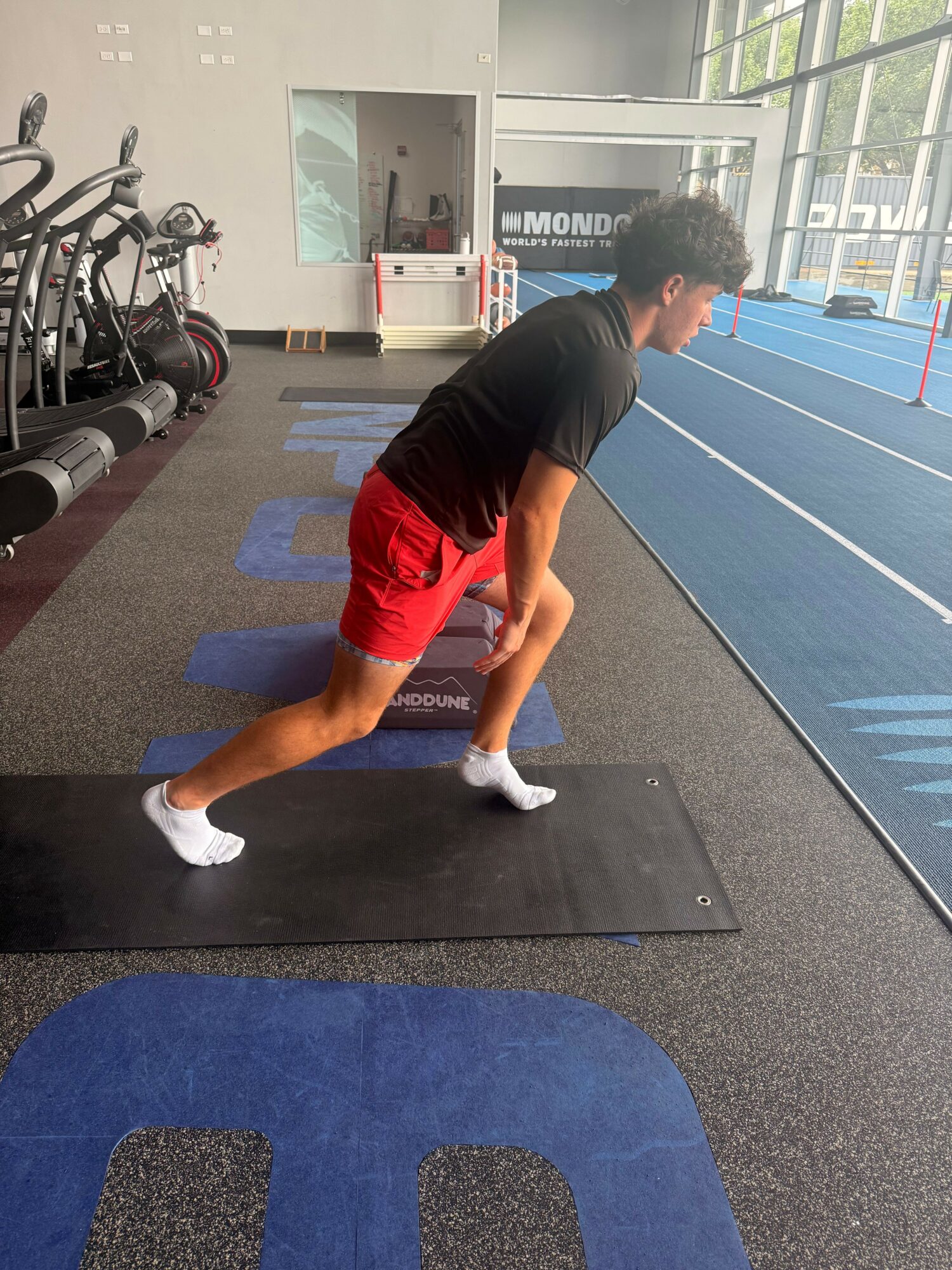 Person stretching on a black mat in a gym, wearing black shirt, red shorts, and white shoes.