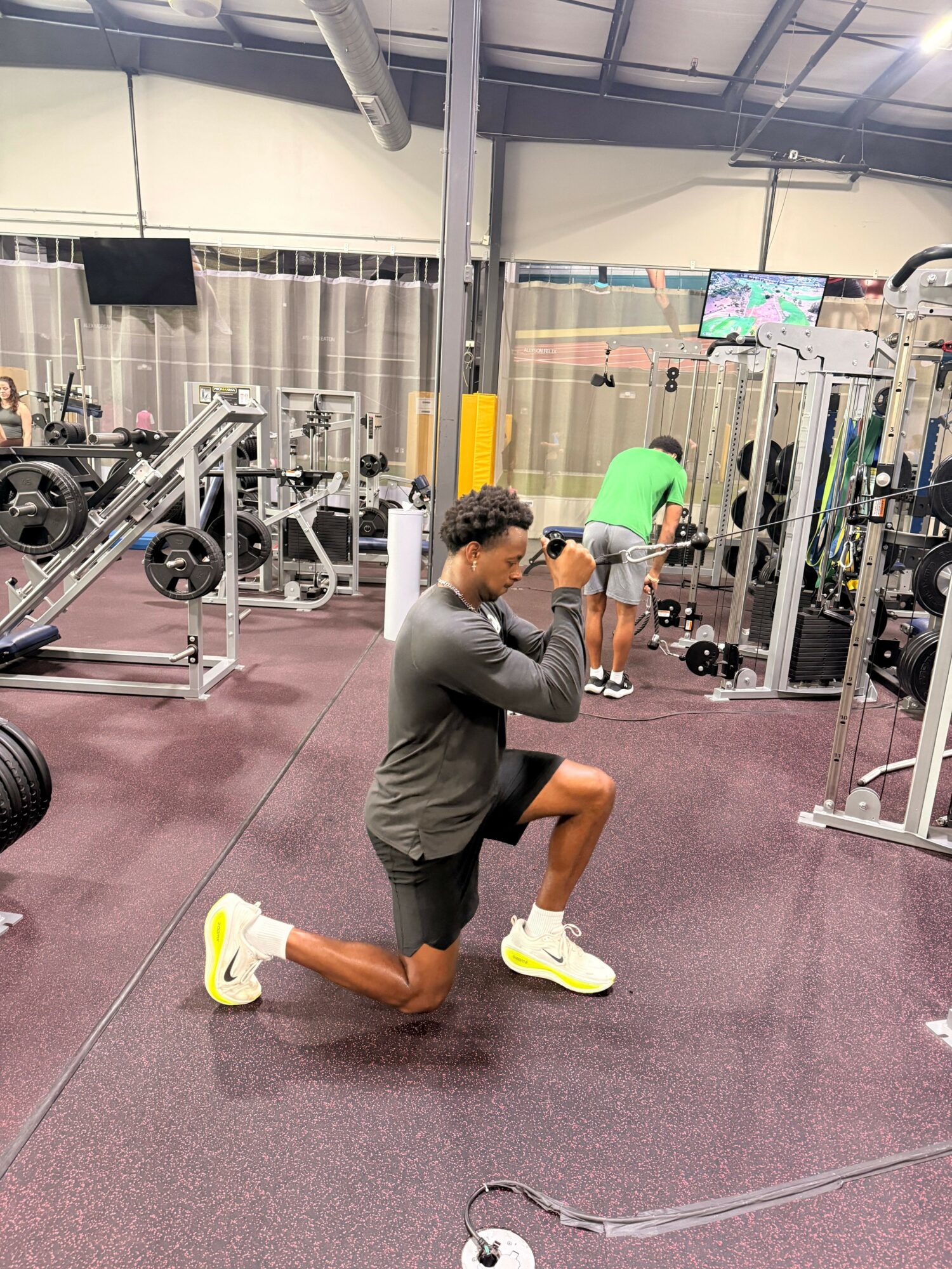 Person in gray athletic clothing doing a stretching exercise in a gym with equipment and other people in the background.