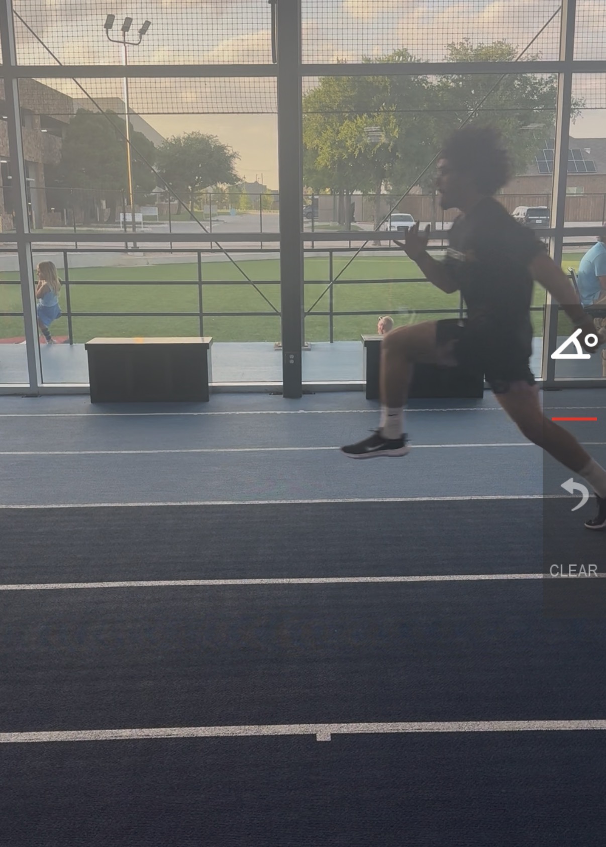 Person running indoors near large windows with a view of a sports field outside, sunlight streaming in.
