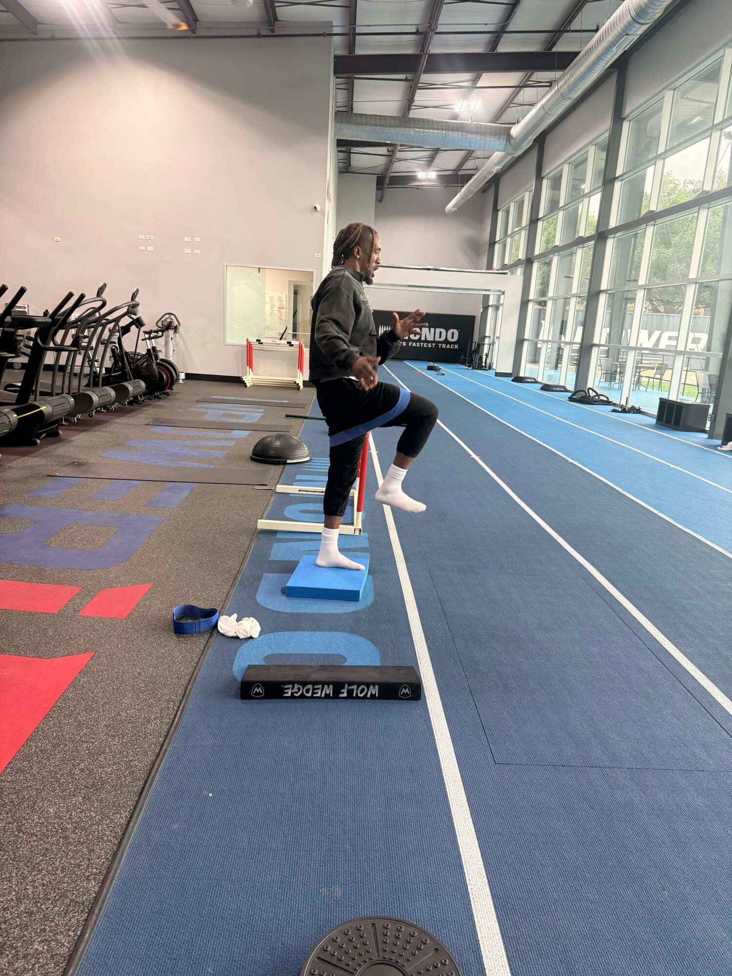 Person jumping over a blue hurdle on an indoor track, with gym equipment and chairs in the background.