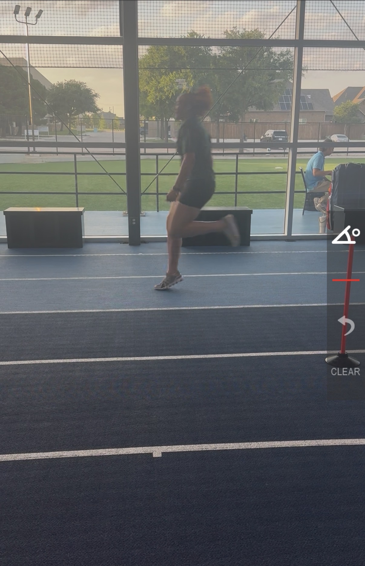 Person standing on a track field inside a glass enclosure, with trees and a person sitting outside visible through the glass.