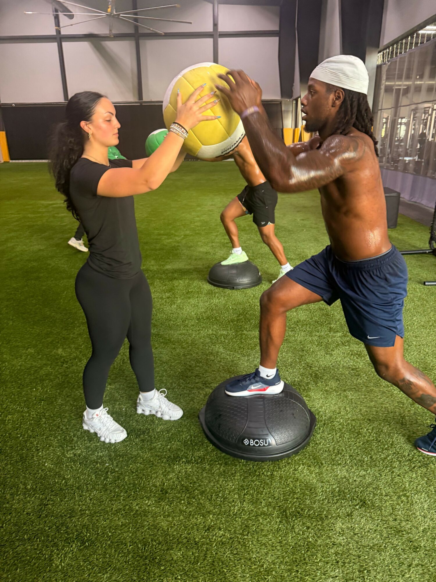 A man and a woman exercise indoors on artificial grass, using a balance board and a medicine ball, with gym equipment in background.