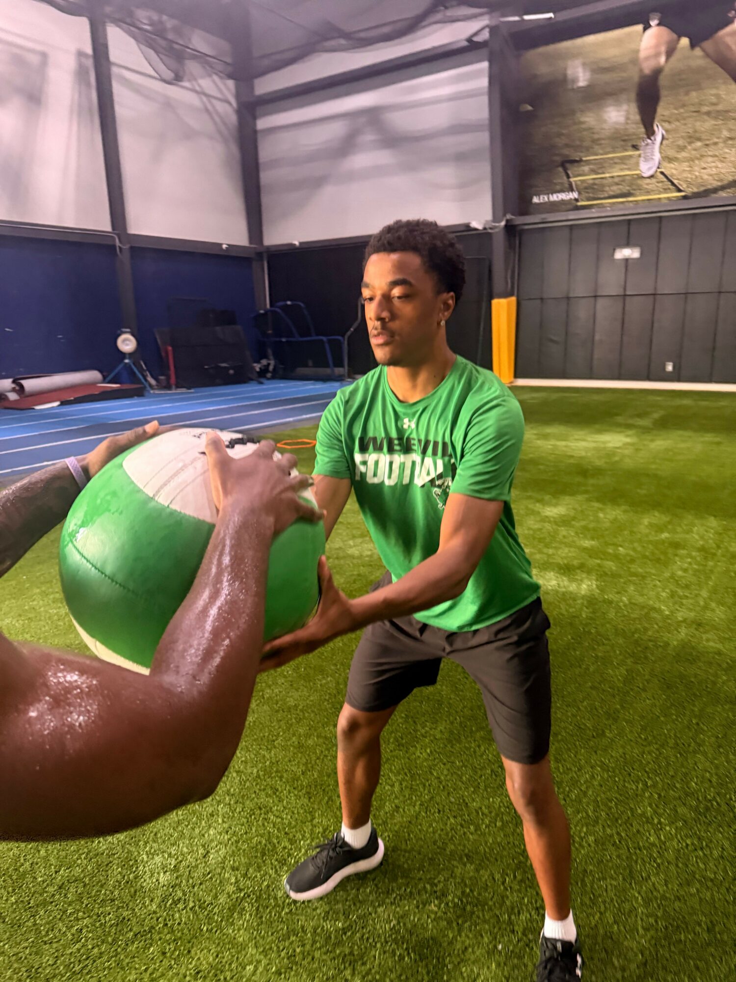 Person in green shirt and black shorts holding a green ball on artificial grass, indoors.