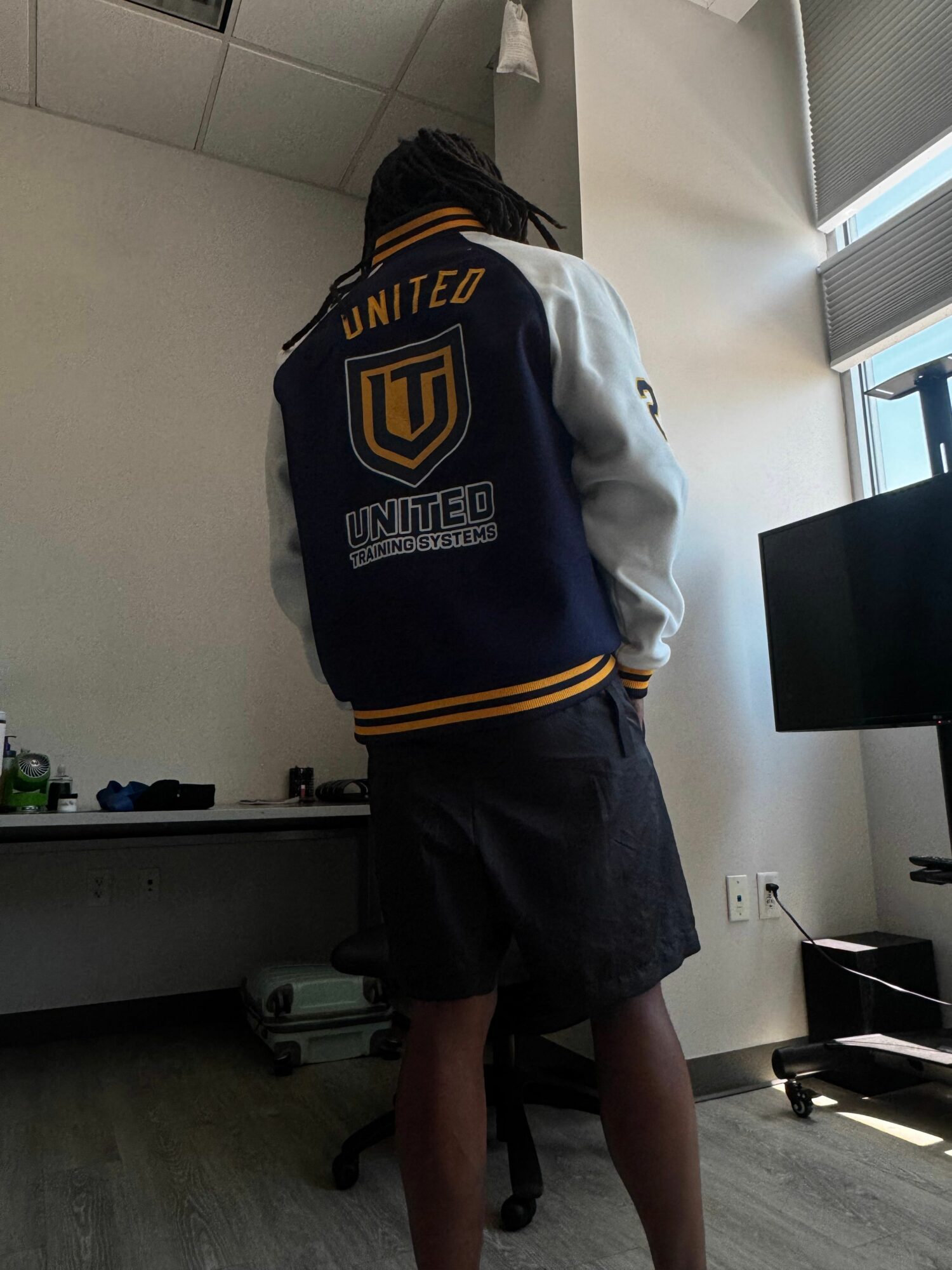 Person standing indoors, wearing a hoodie and shorts, facing away from the camera, with a window and furniture in the background.