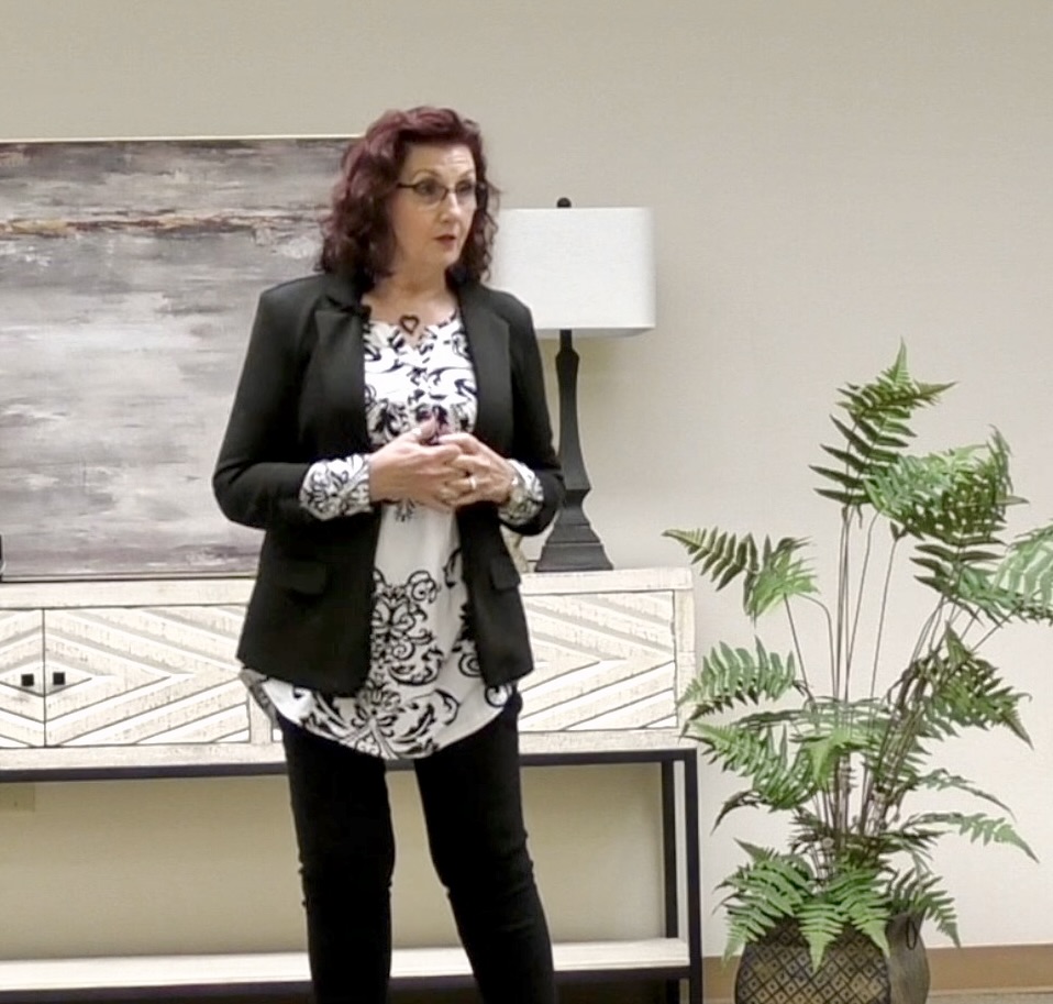 Woman with curly hair wearing a black blazer and patterned top, standing indoors near a potted plant and a lamp.