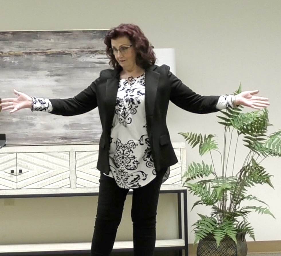 Woman with curly hair and glasses stands with arms outstretched indoors, next to a potted plant and abstract artwork.