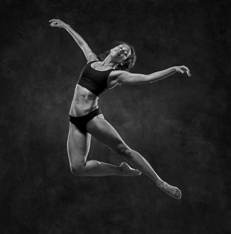 Ballet dancer mid-air with arms and legs extended, wearing a black sports bra and shorts, against a dark background.