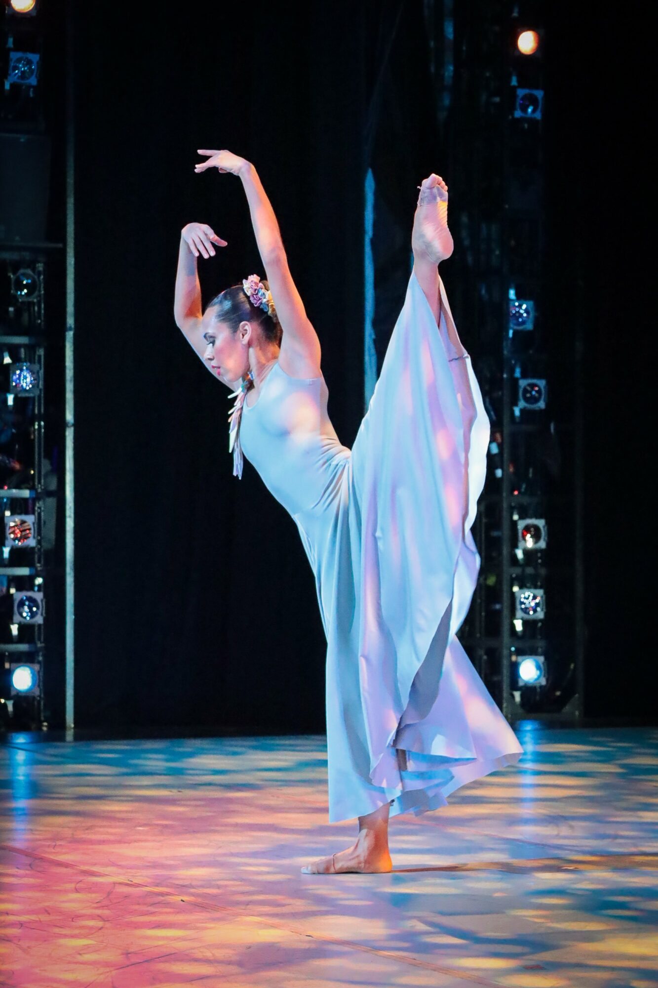 A dancer performs a pose on stage with one leg raised and arms curved above her head, wearing a flowing dress.
