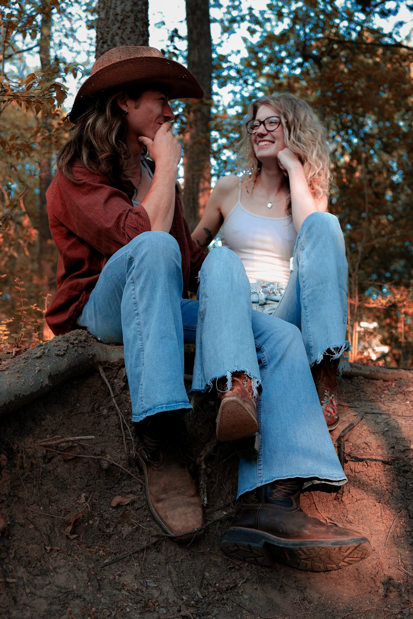 Two women sitting on a tree root outdoors, smiling and talking, surrounded by autumn trees.