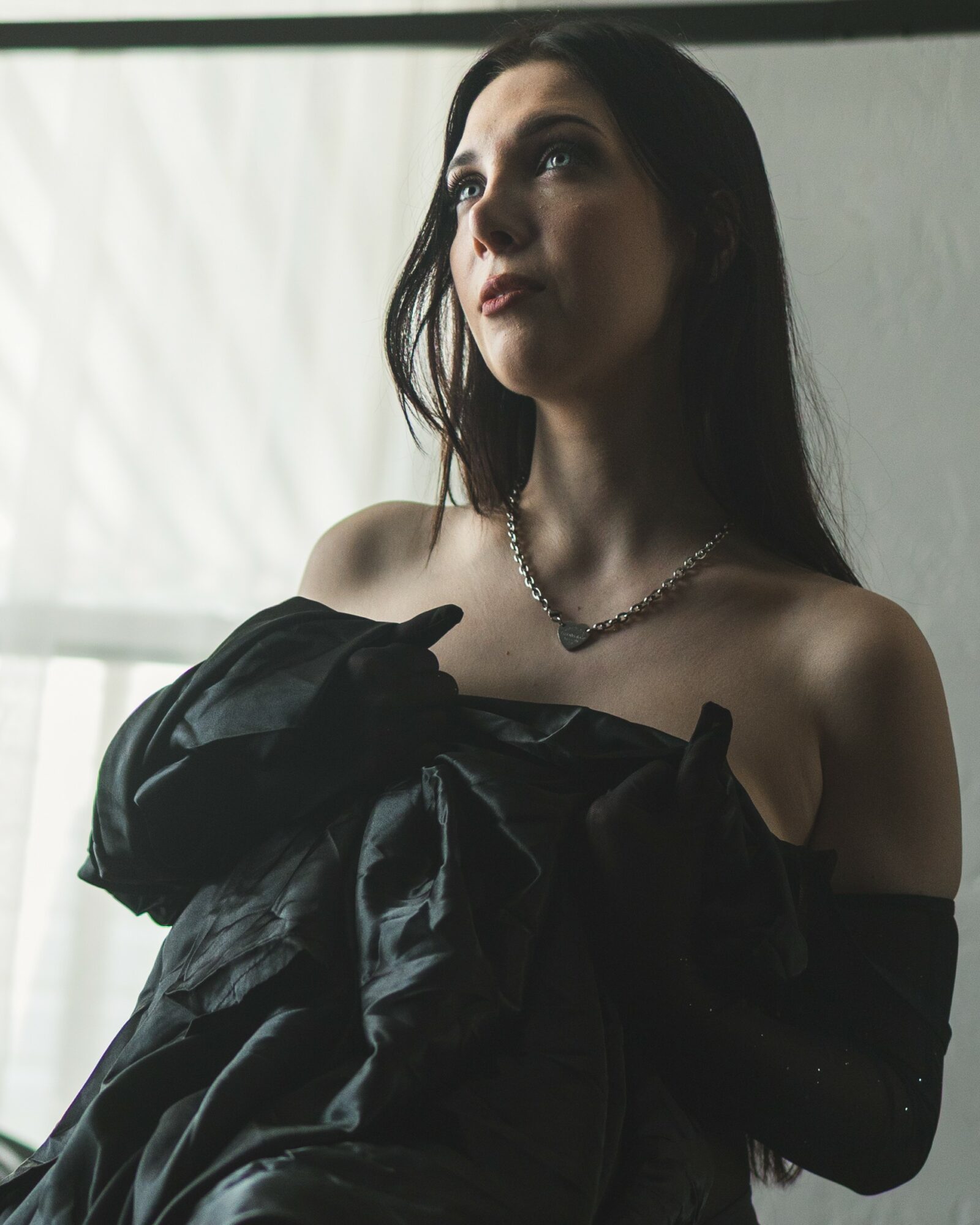 Woman with dark hair wearing a black off-shoulder dress and necklace, looking to the side indoors.
