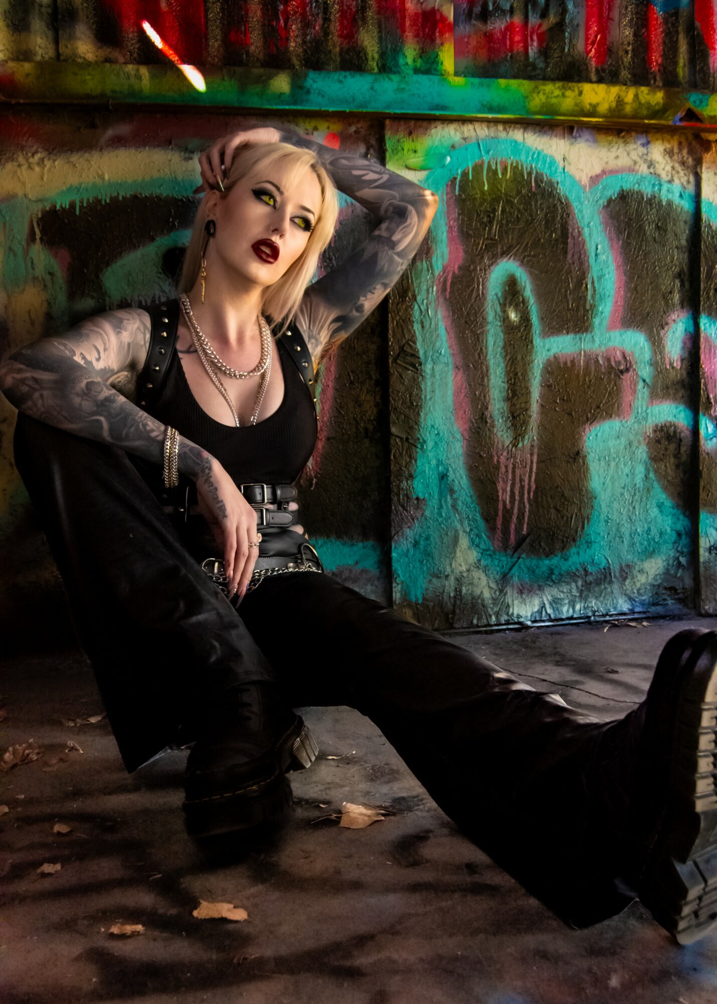 Woman with platinum blonde hair and tattoos, sitting on the ground against a graffiti-covered wall, wearing dark clothing and jewelry.