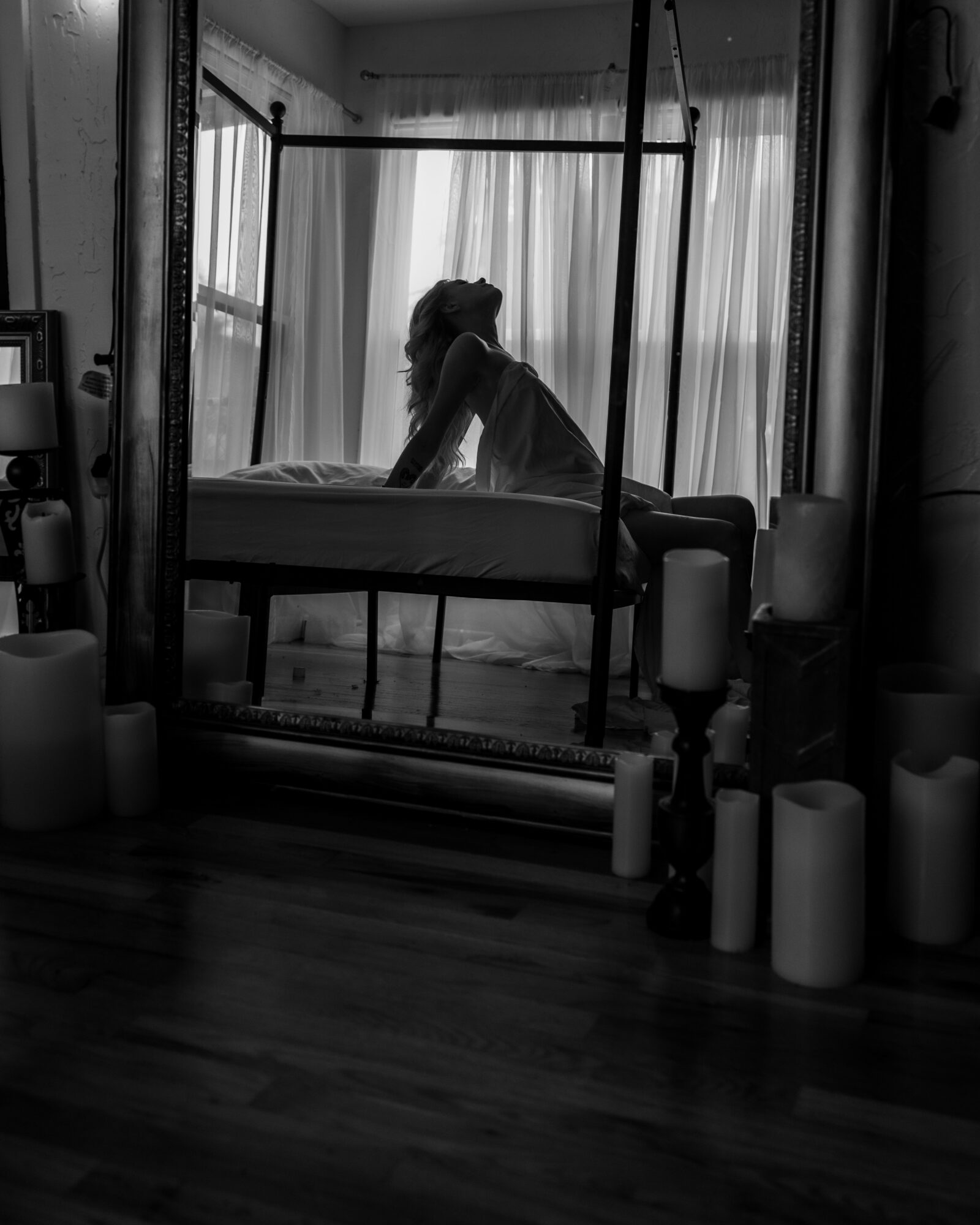 Person sitting on a bed near a window with curtains, viewed through a mirror, in black and white.