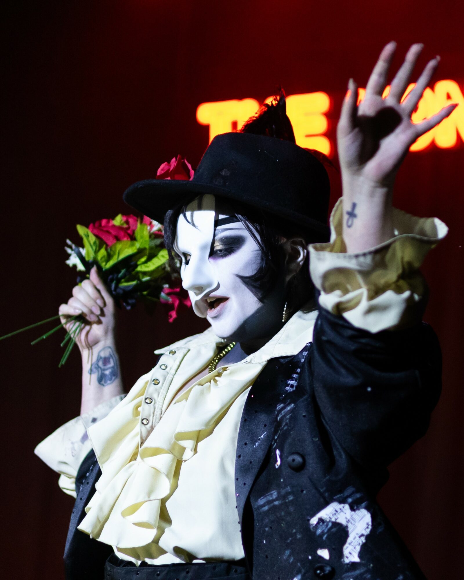 Performer with face paint, black hat, and costume holding a bouquet of flowers, raising one hand, in front of a red background.