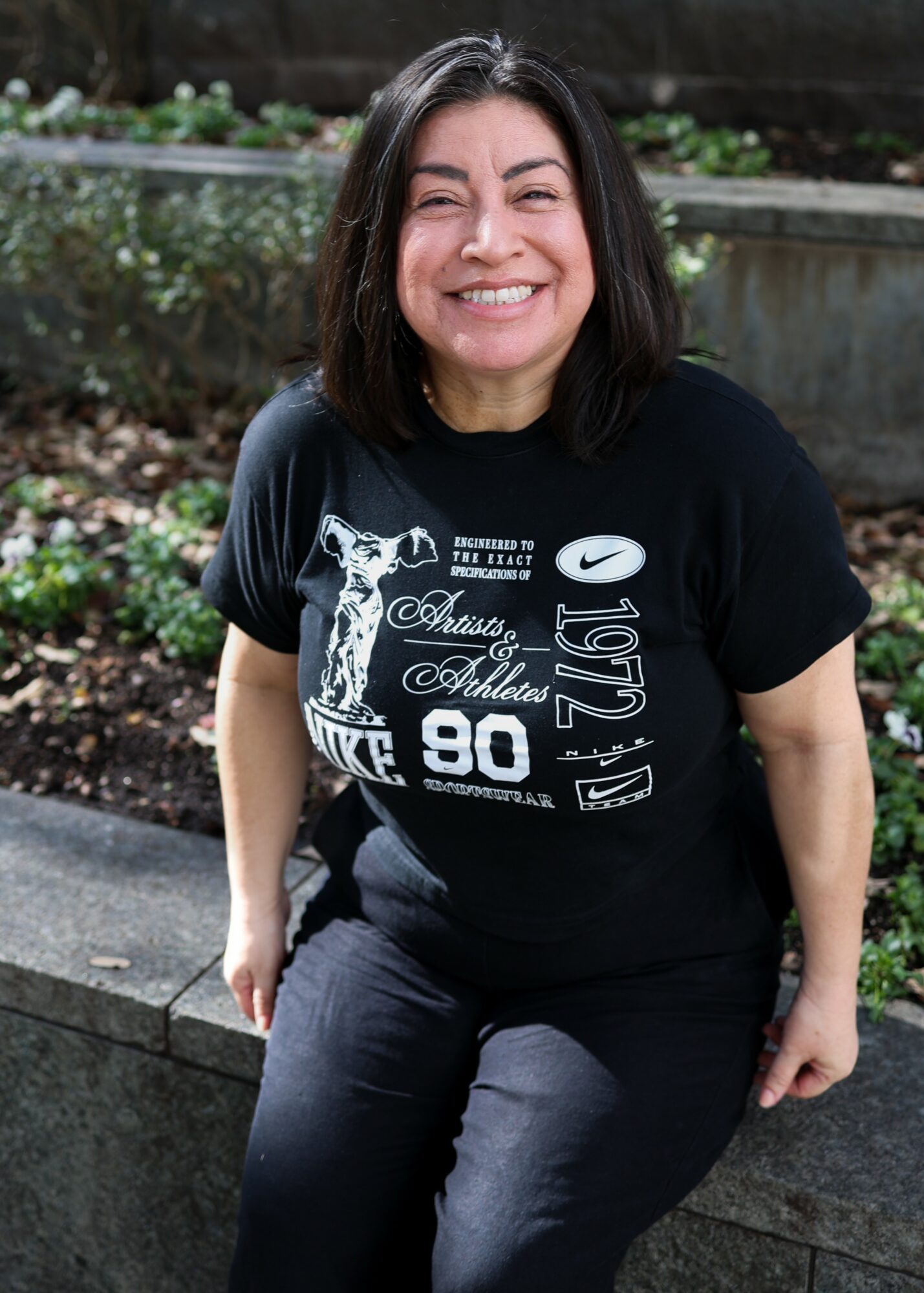 Smiling woman with shoulder-length dark hair sitting outdoors on a stone ledge, wearing a black graphic T-shirt and dark pants.