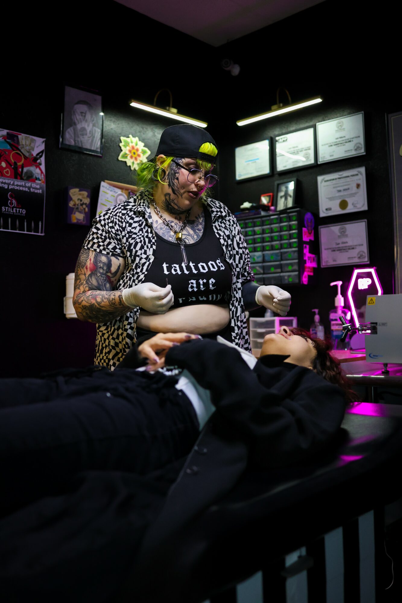 Tattoo artist with colorful clothing and accessories working on a client in a dimly lit studio with certificates on the wall.