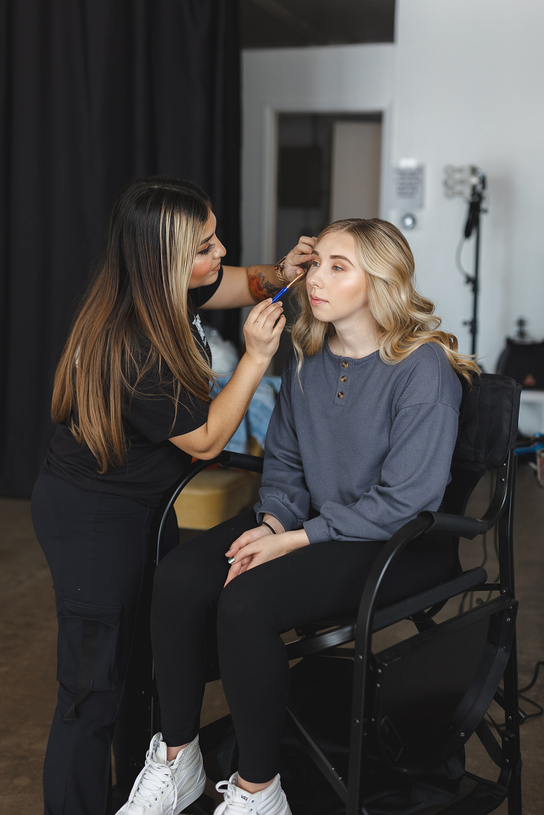 Makeup artist applying makeup to a seated woman with blonde hair in a studio setting.