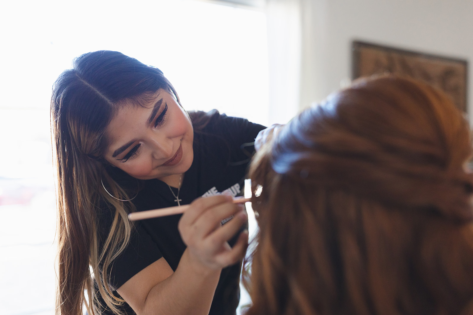 Woman applying makeup to another person's face indoors, with a window and framed picture in the background.