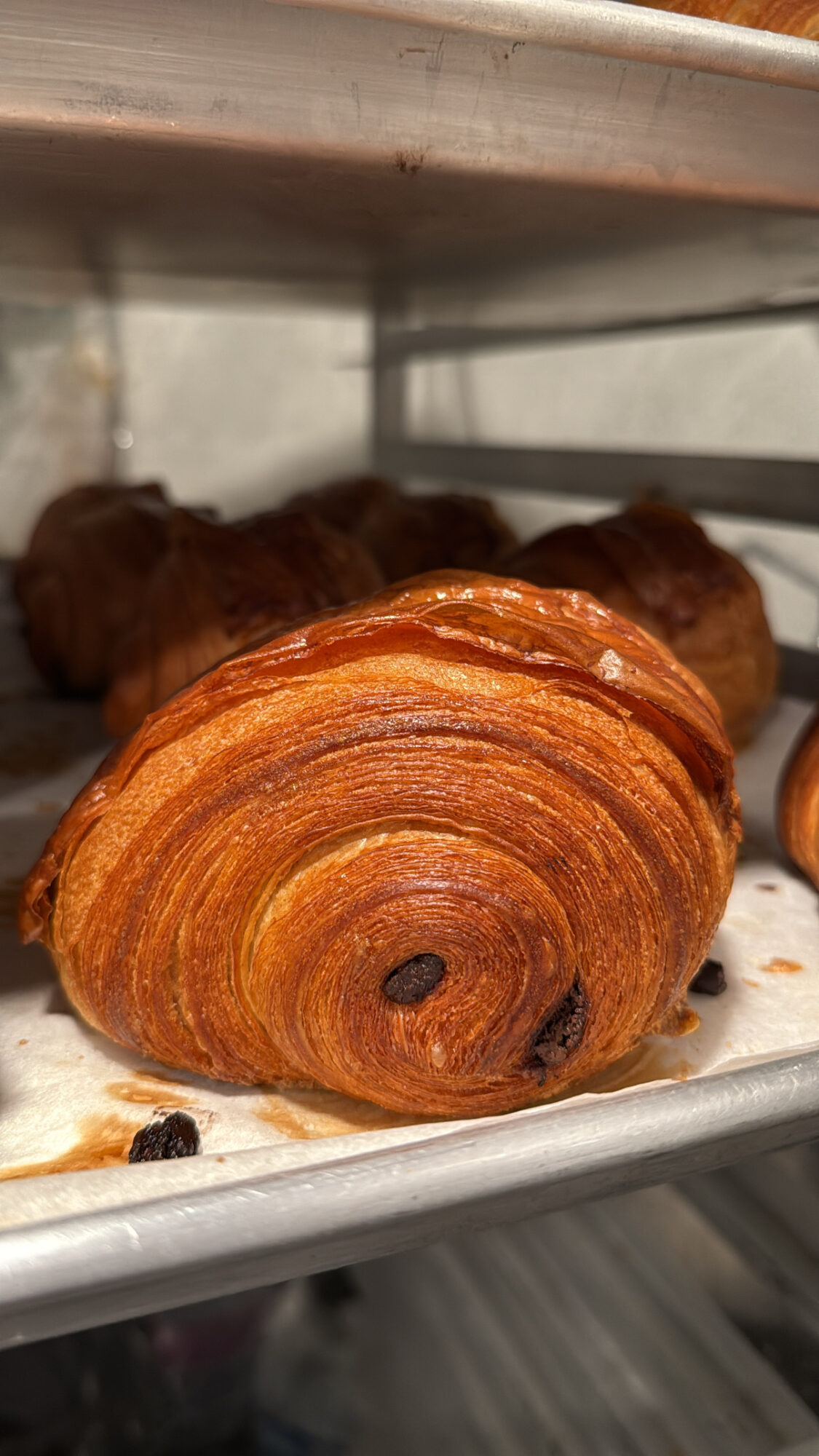 Close-up of a croissant with a golden-brown, flaky exterior on a tray, with other baked goods in the background.