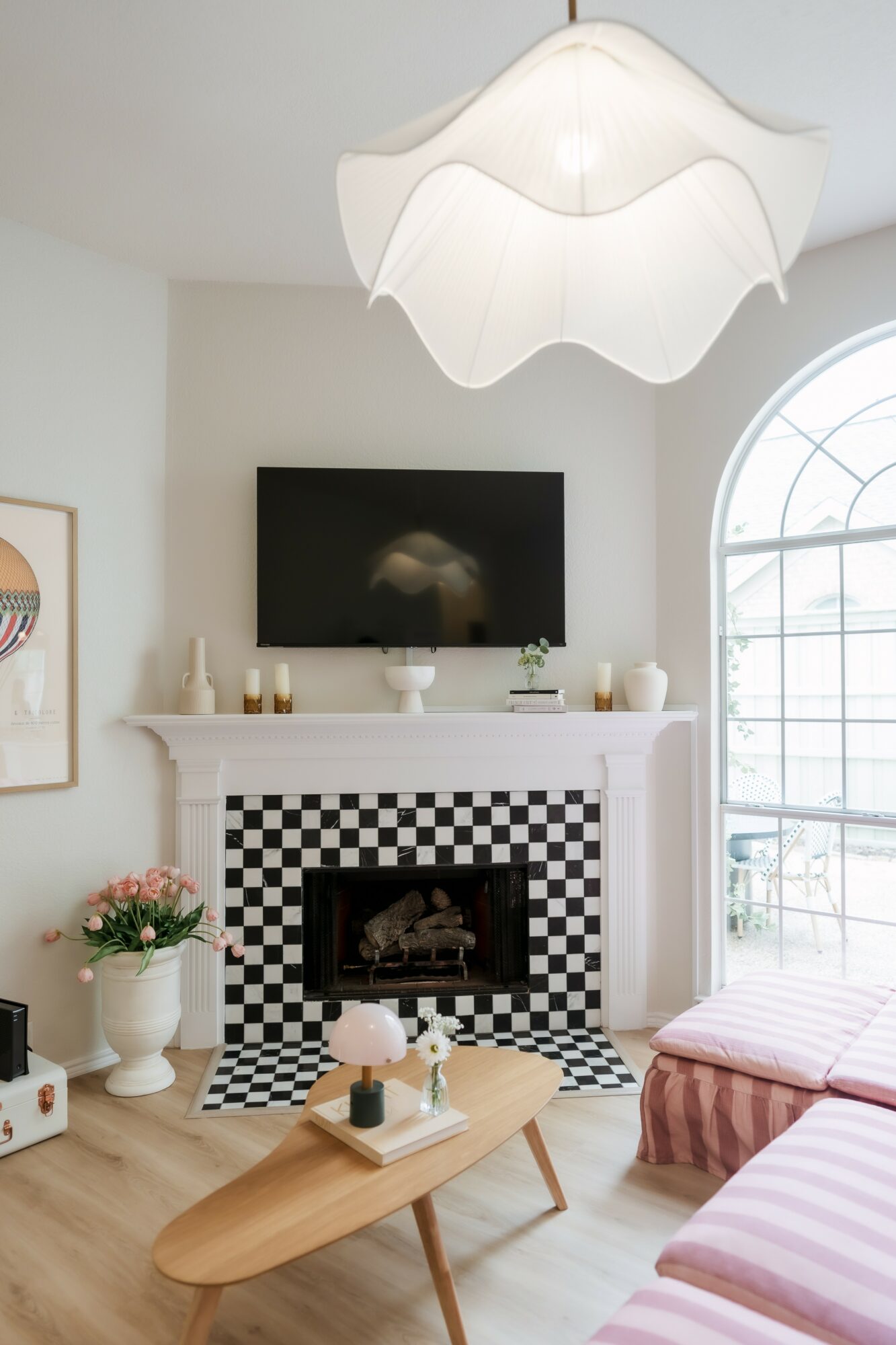 Living room with fireplace, wall-mounted TV, pink striped sofa, and a wooden coffee table, bright natural light from large window.