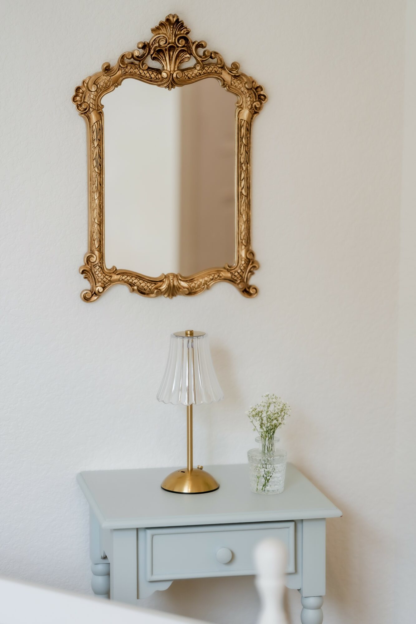 Decorative gold-framed mirror hangs above a white table with a gold lamp and a small vase of flowers.