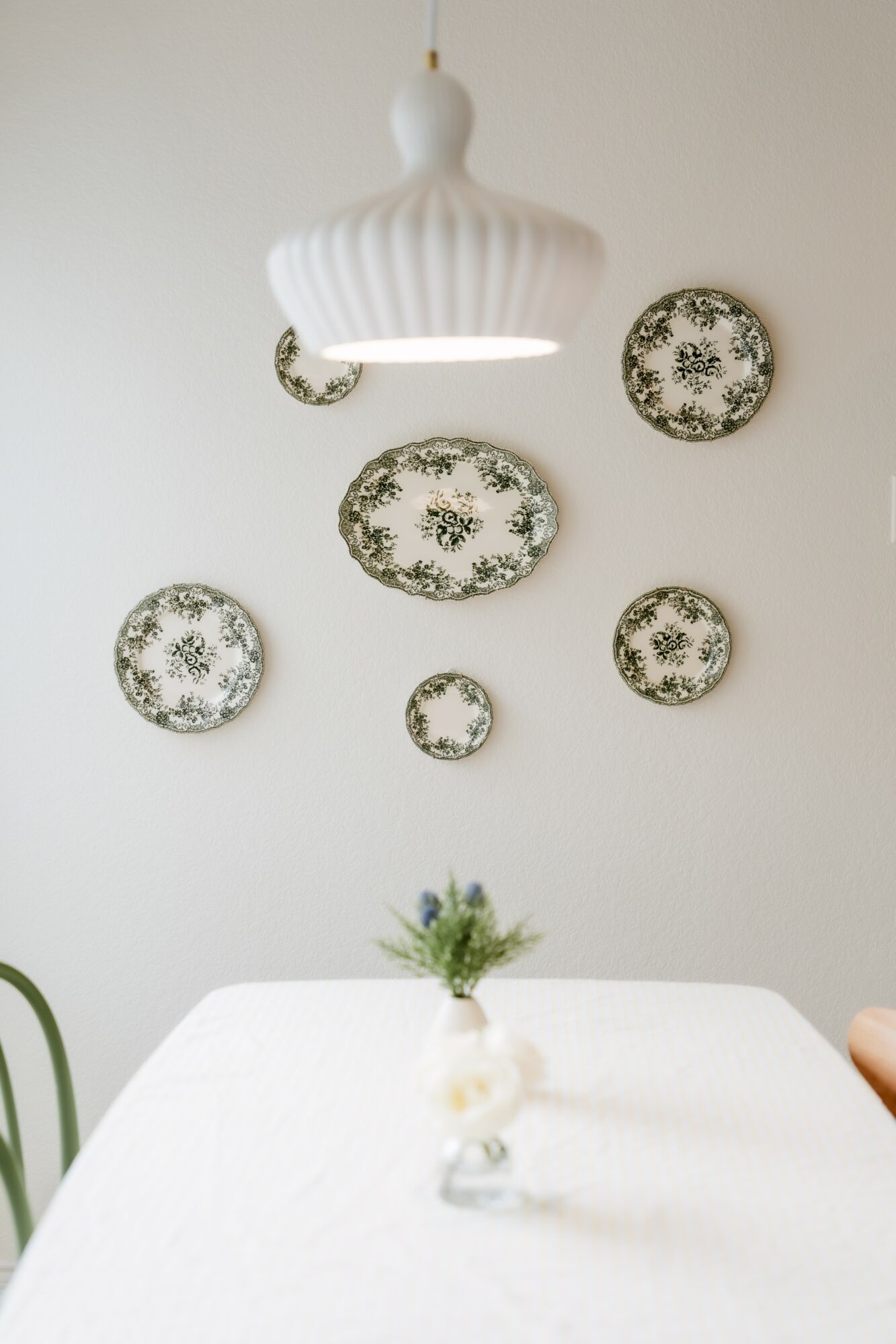 White pendant lamp hangs above a white table with a small plant in a glass vase, decorated with patterned plates on the wall.