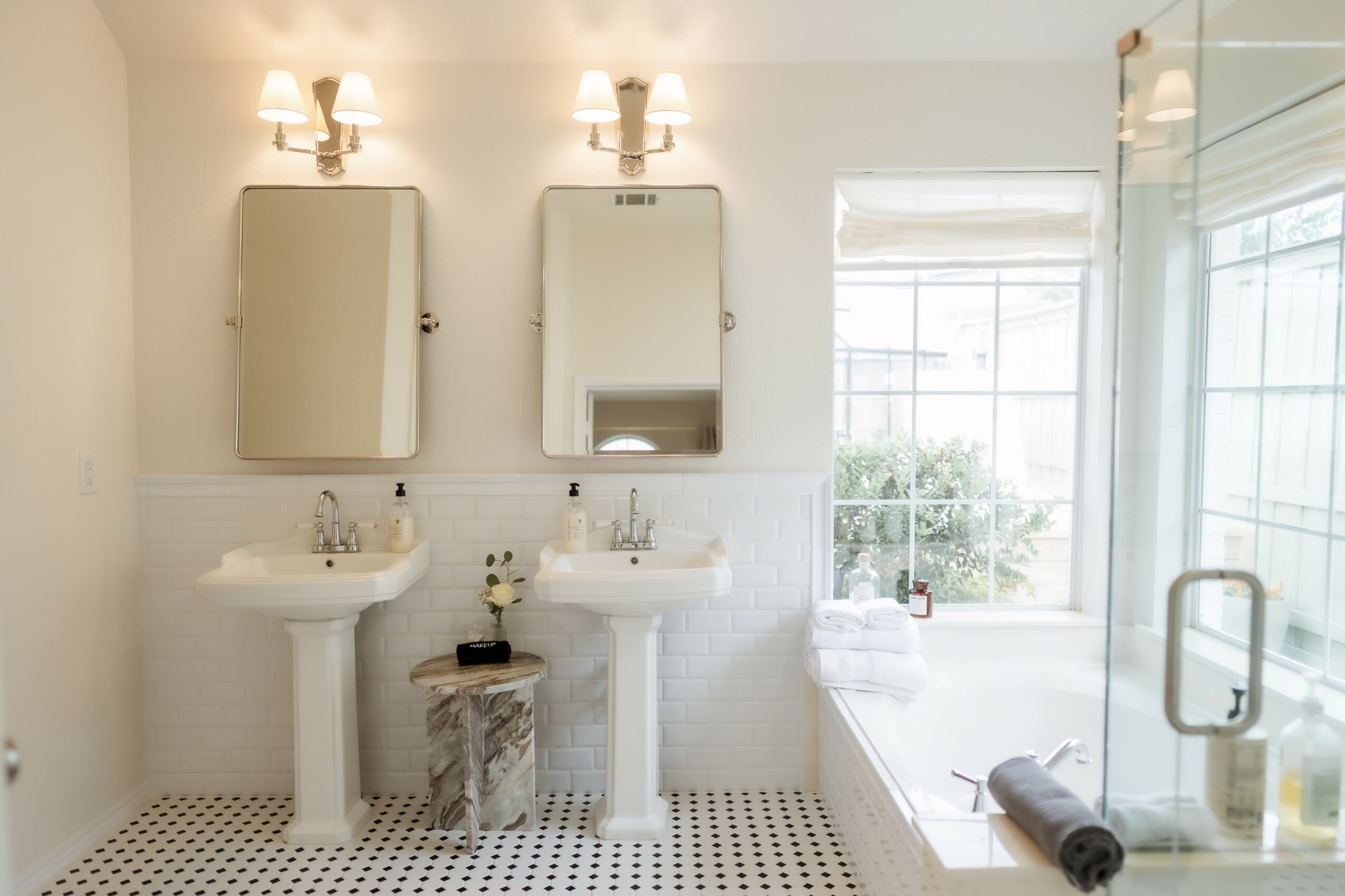 Bathroom with two sinks, mirrors, and a window with a view outside, featuring a bathtub and a rolled towel.