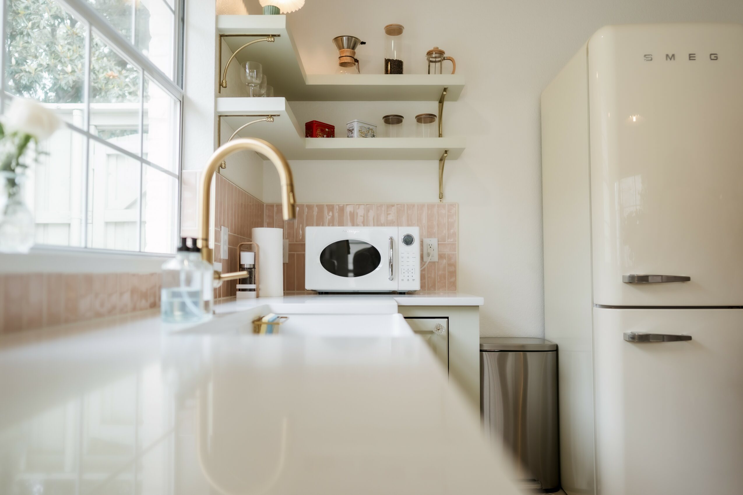 Kitchen with white cabinets, open shelves, microwave, and a refrigerator, illuminated by natural light from a window.