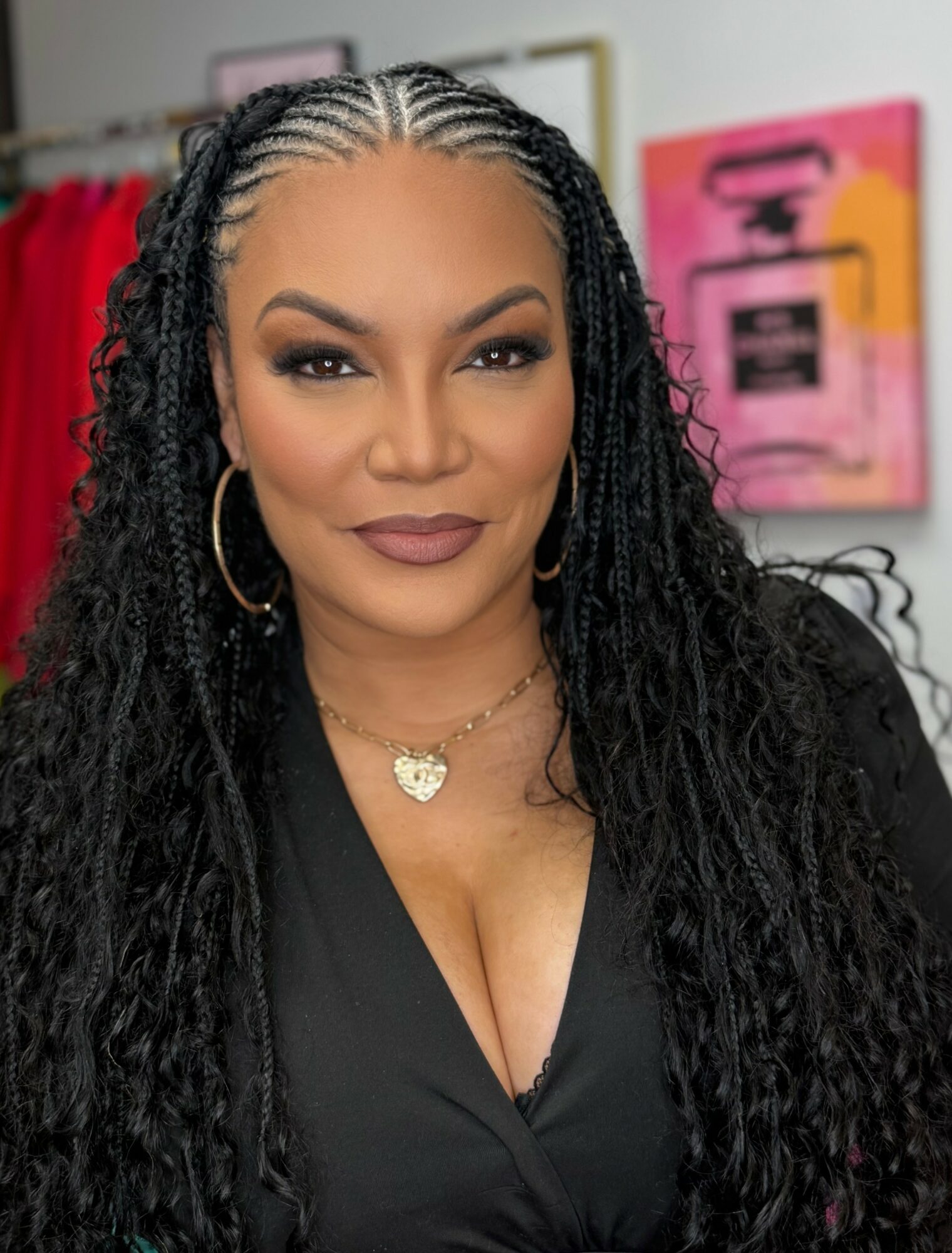 Woman with braided hair, wearing hoop earrings and a necklace, smiling in front of colorful artwork.