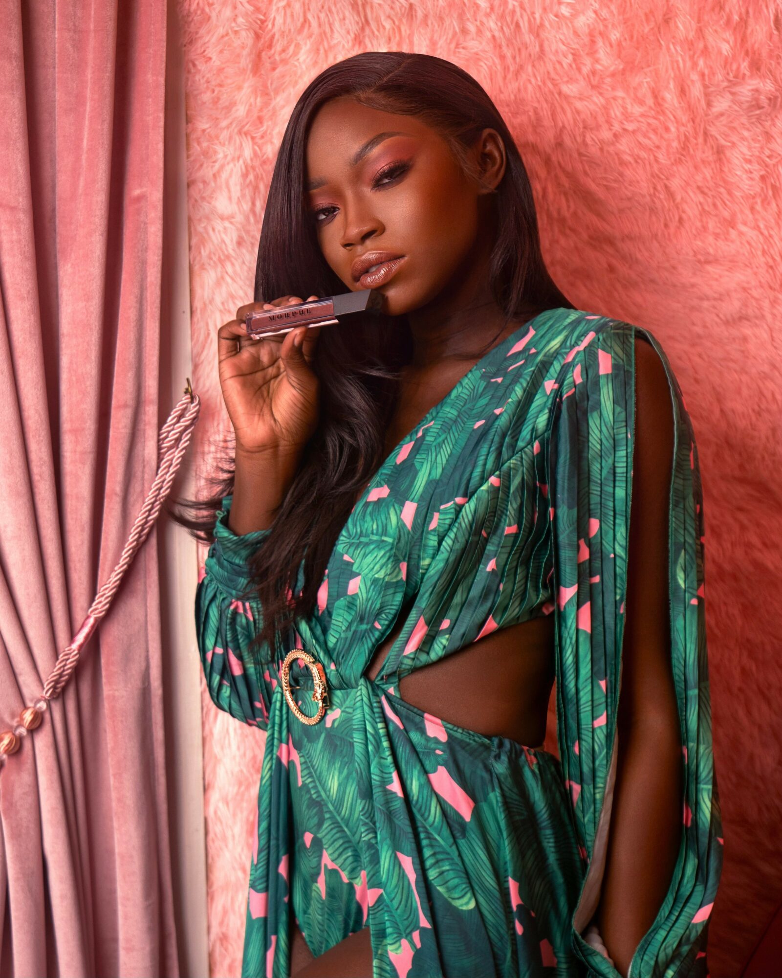 Woman with long black hair holding a gun, wearing a green patterned dress with cutouts, standing against pink textured background.
