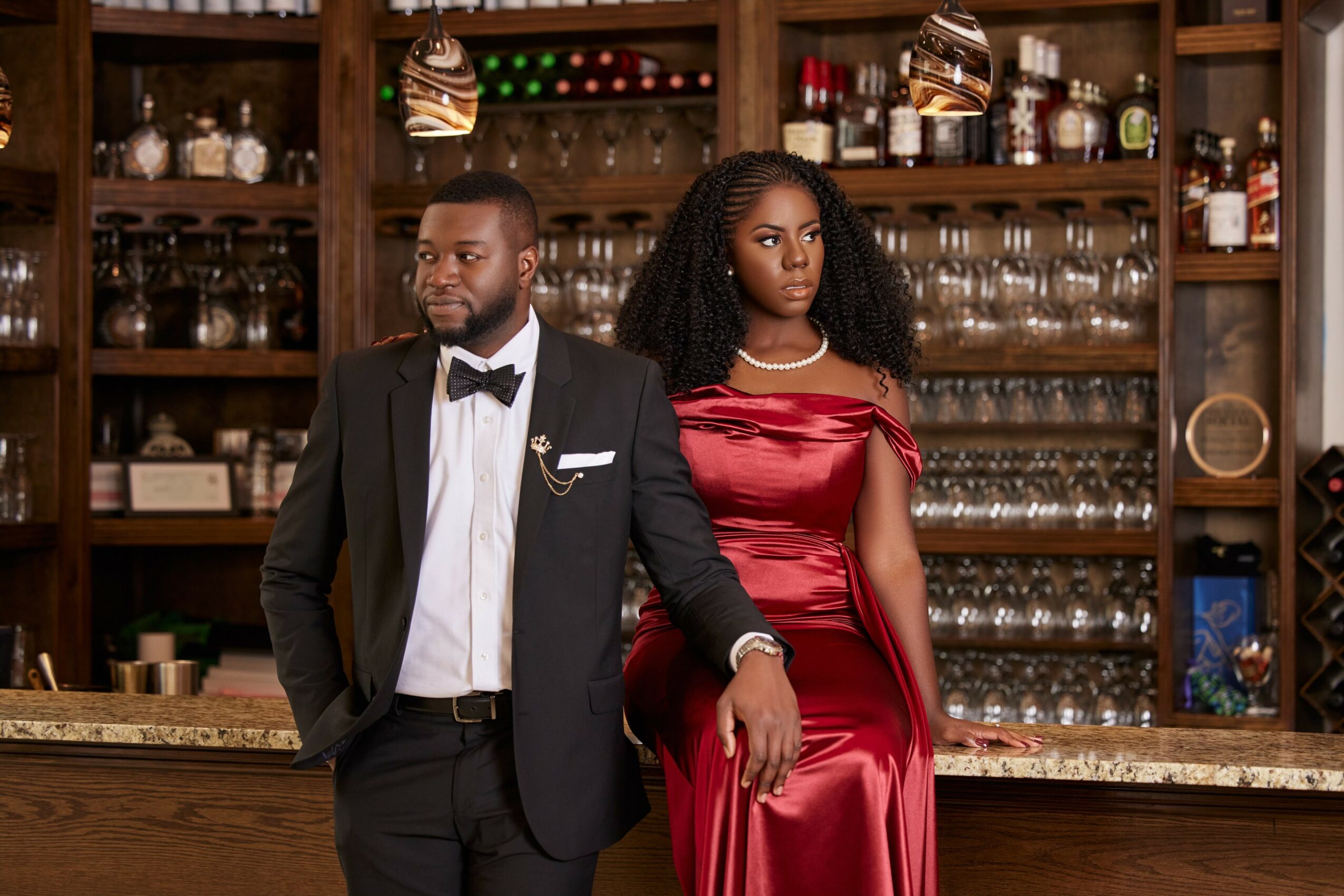 A man and woman dressed formally in a bar or restaurant setting with shelves of glasses and bottles behind them.