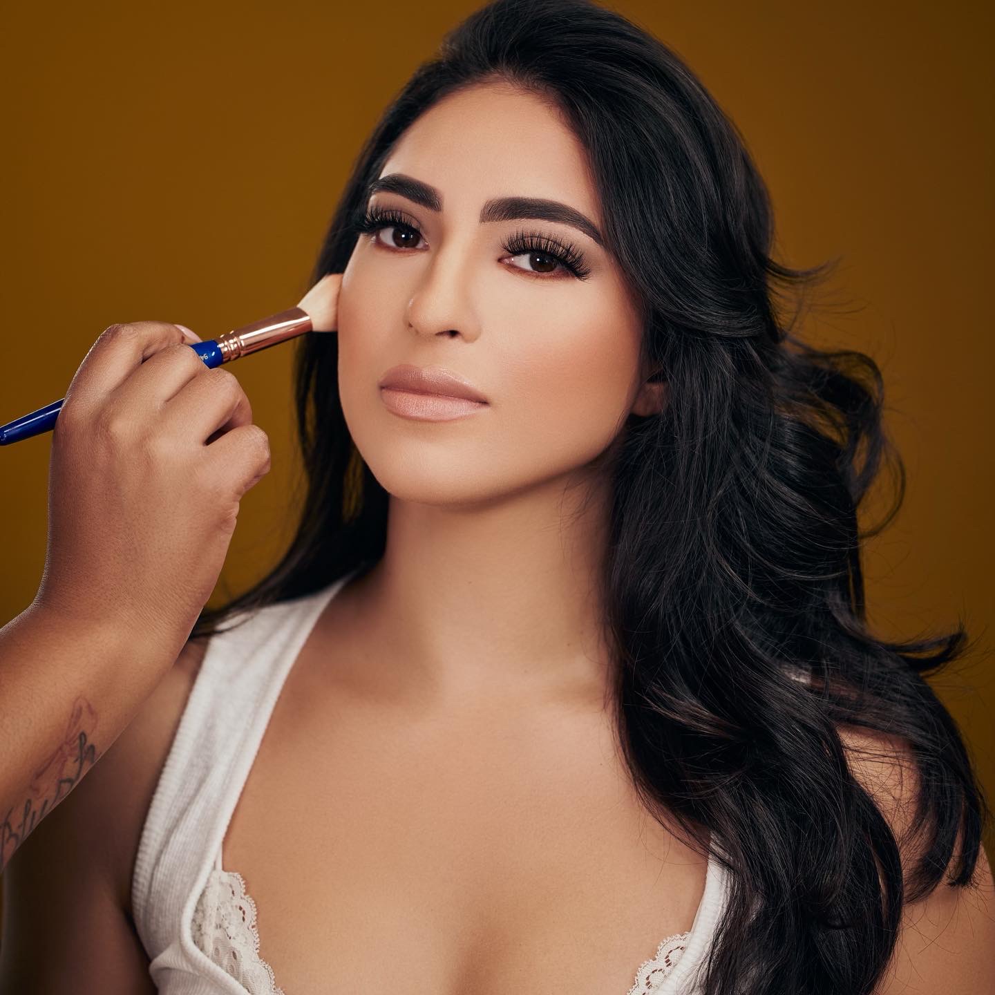 Woman with long dark hair and makeup, being styled with a brush, against a brown background.