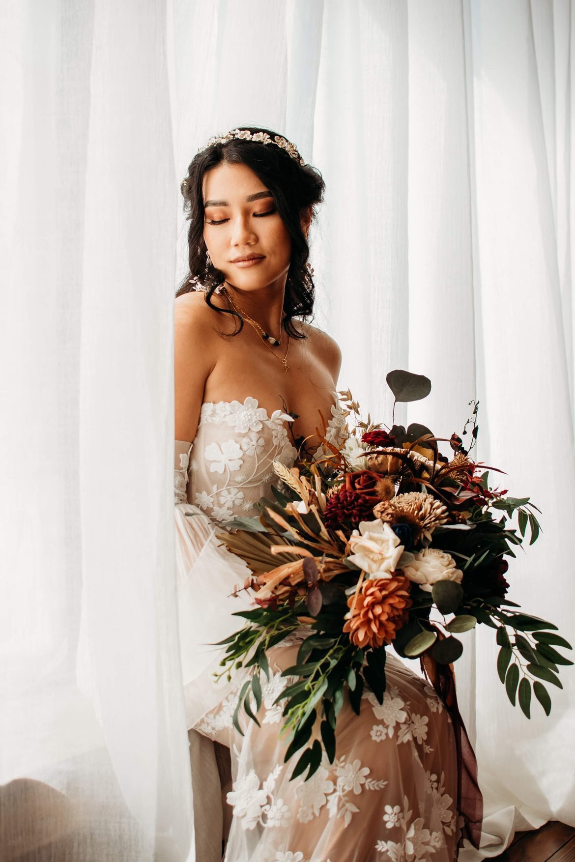 Woman in wedding dress holding a large bouquet, standing near white curtains, eyes closed, serene expression.